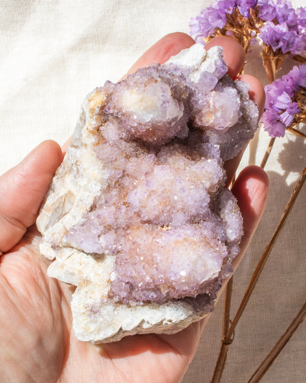 Hand holding a large crystal cluster with purple flowers in the background
