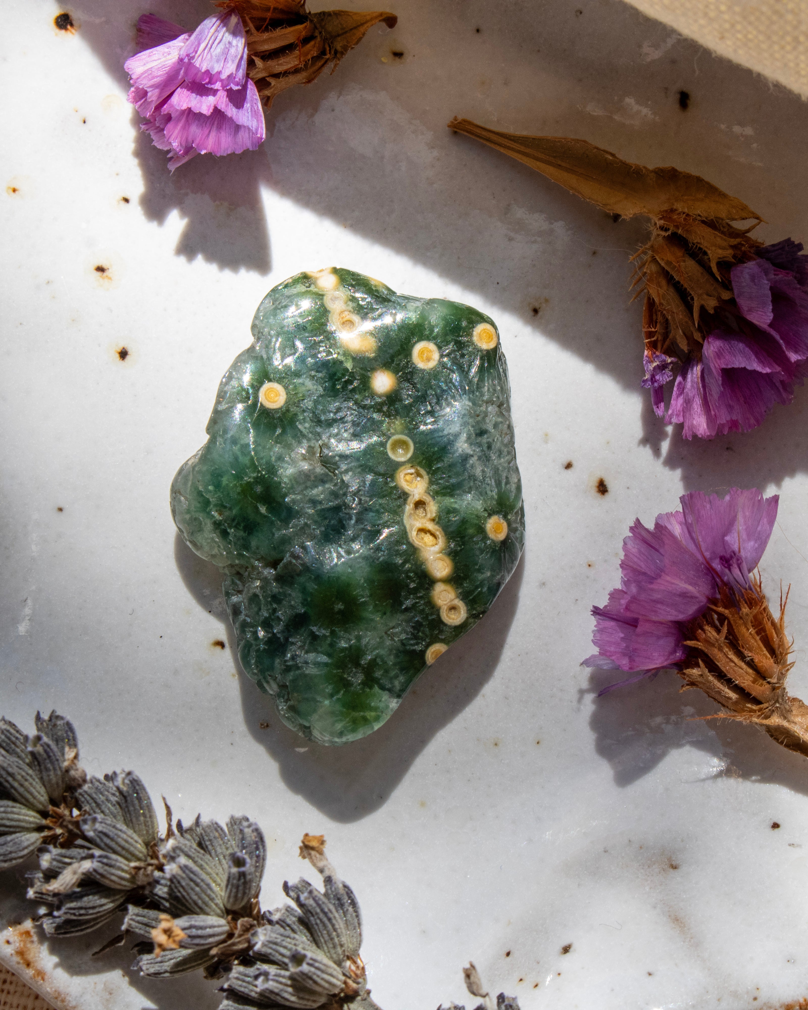 Green crystal rock with yellow crystals on a white surface with purple and brown flowers.