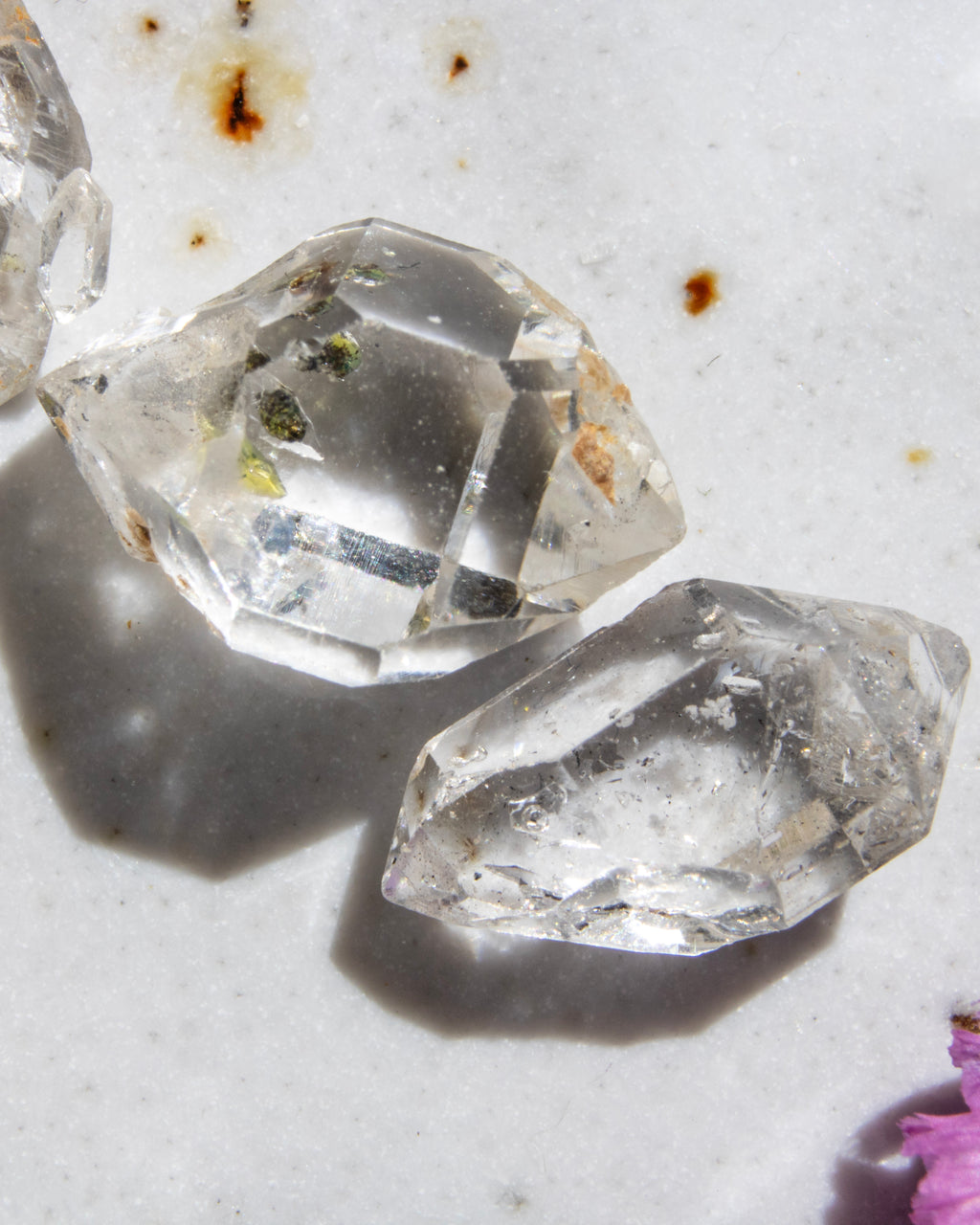 Two clear crystal-like stones on a textured surface with a pink flower at the bottom right corner.
