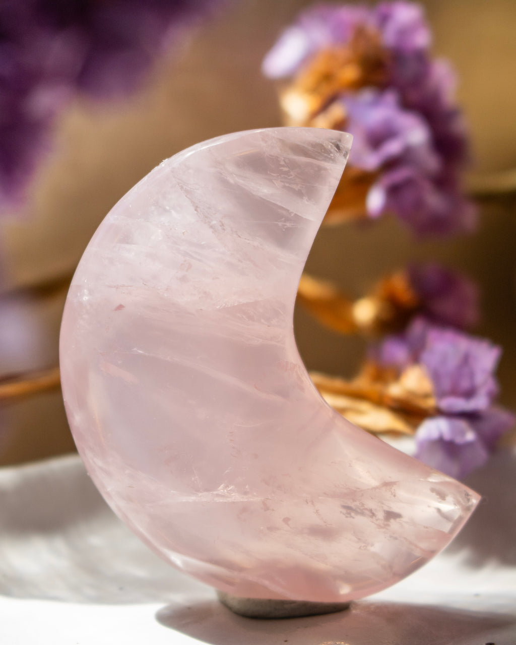 Pink crystal moon shape on a white surface with blurred purple flowers in the background