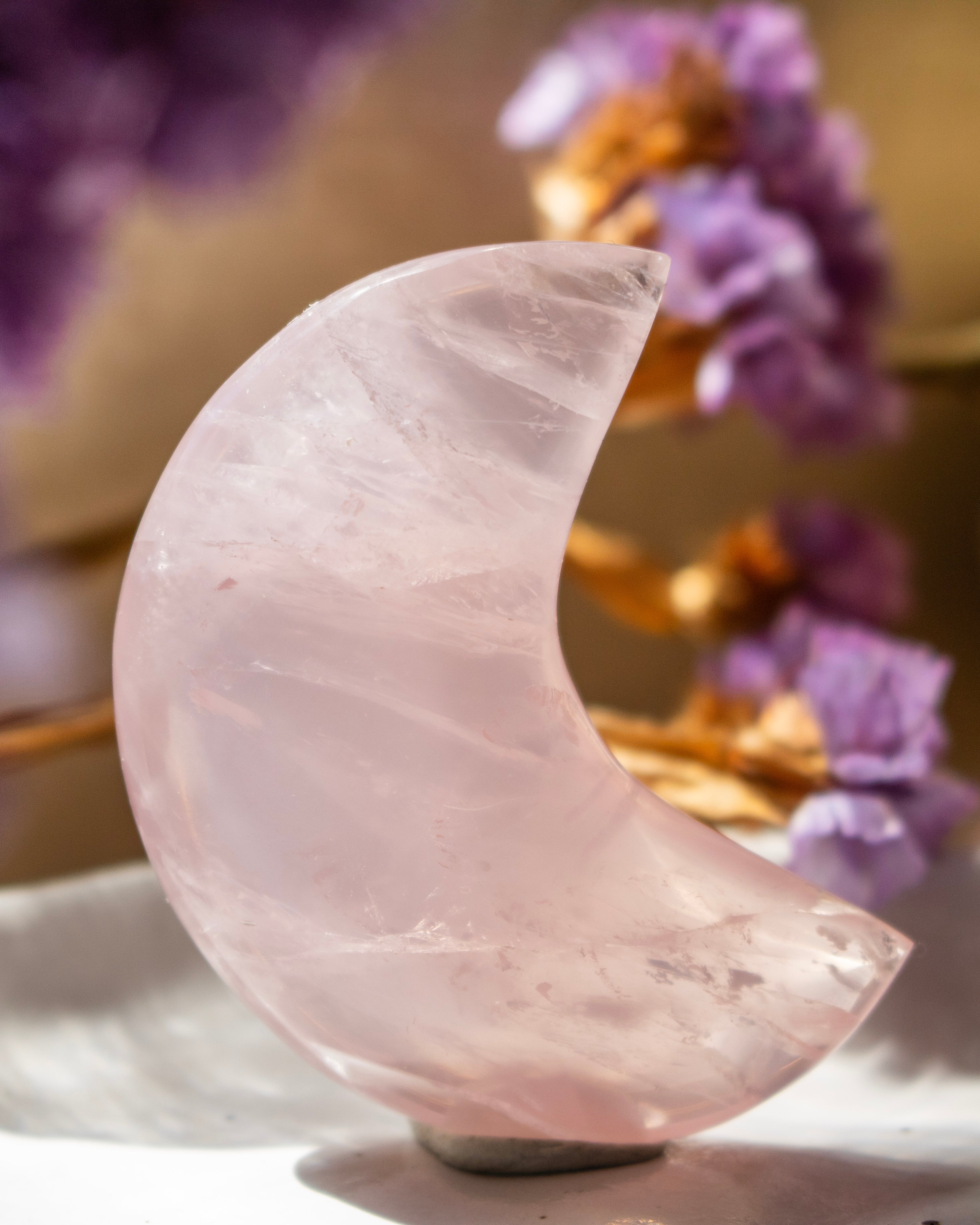 Pink crystal moon shape on a white surface with blurred purple flowers in the background
