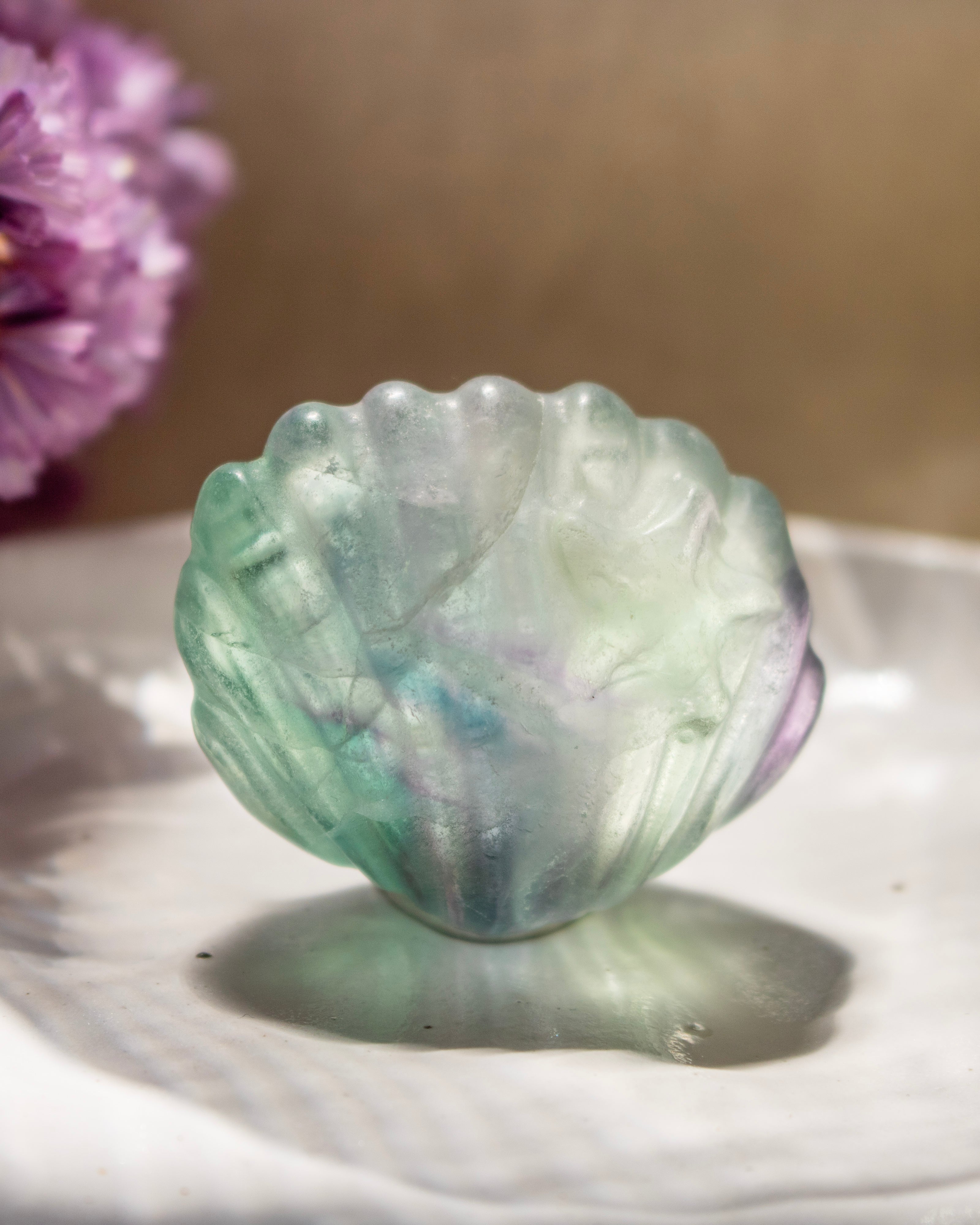 Translucent green stone shaped like a shell on a reflective surface with a blurred purple flower in the background.
