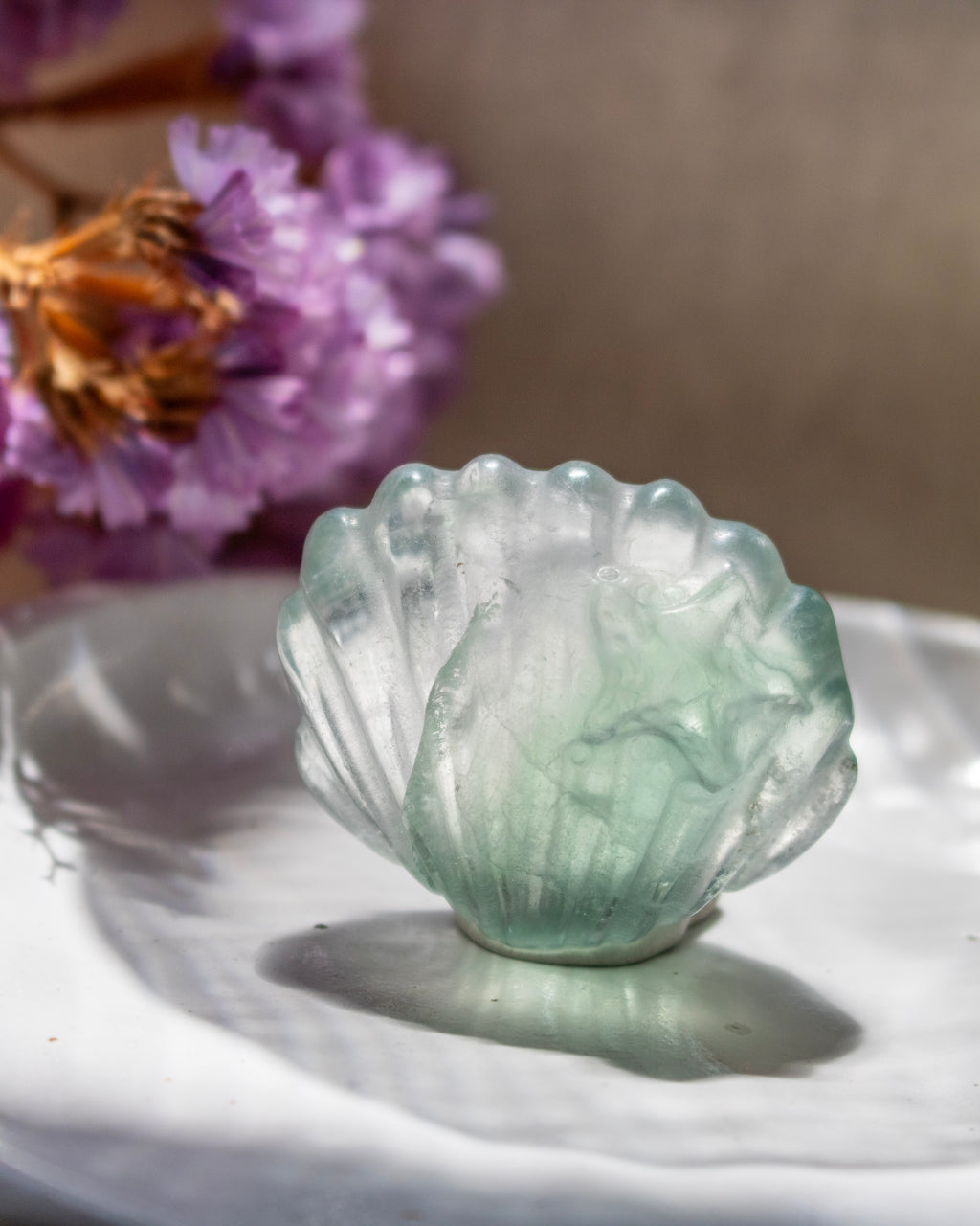 Green crystal shell-shaped object on a white surface with purple flowers in the background