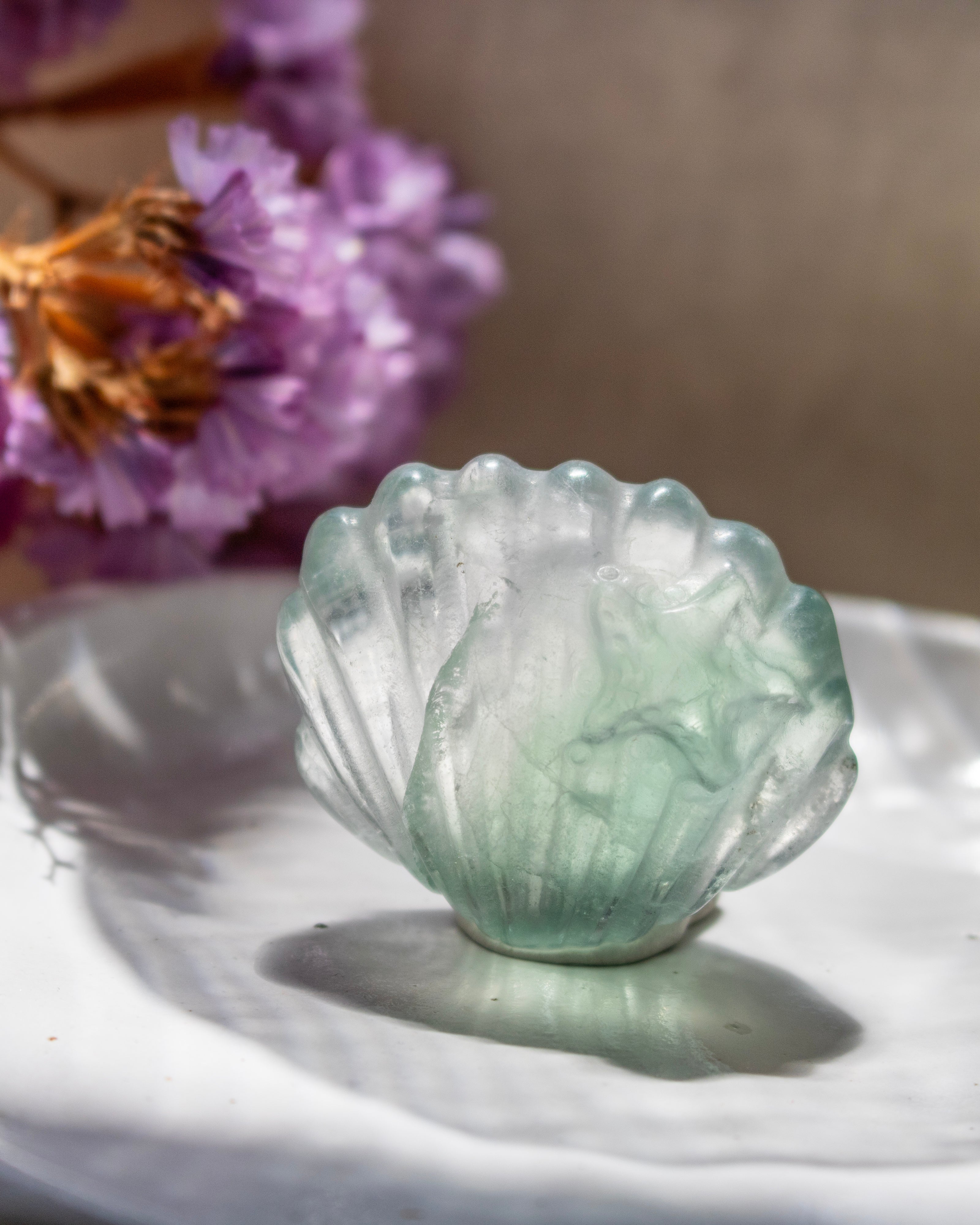 Green crystal shell-shaped object on a white surface with purple flowers in the background