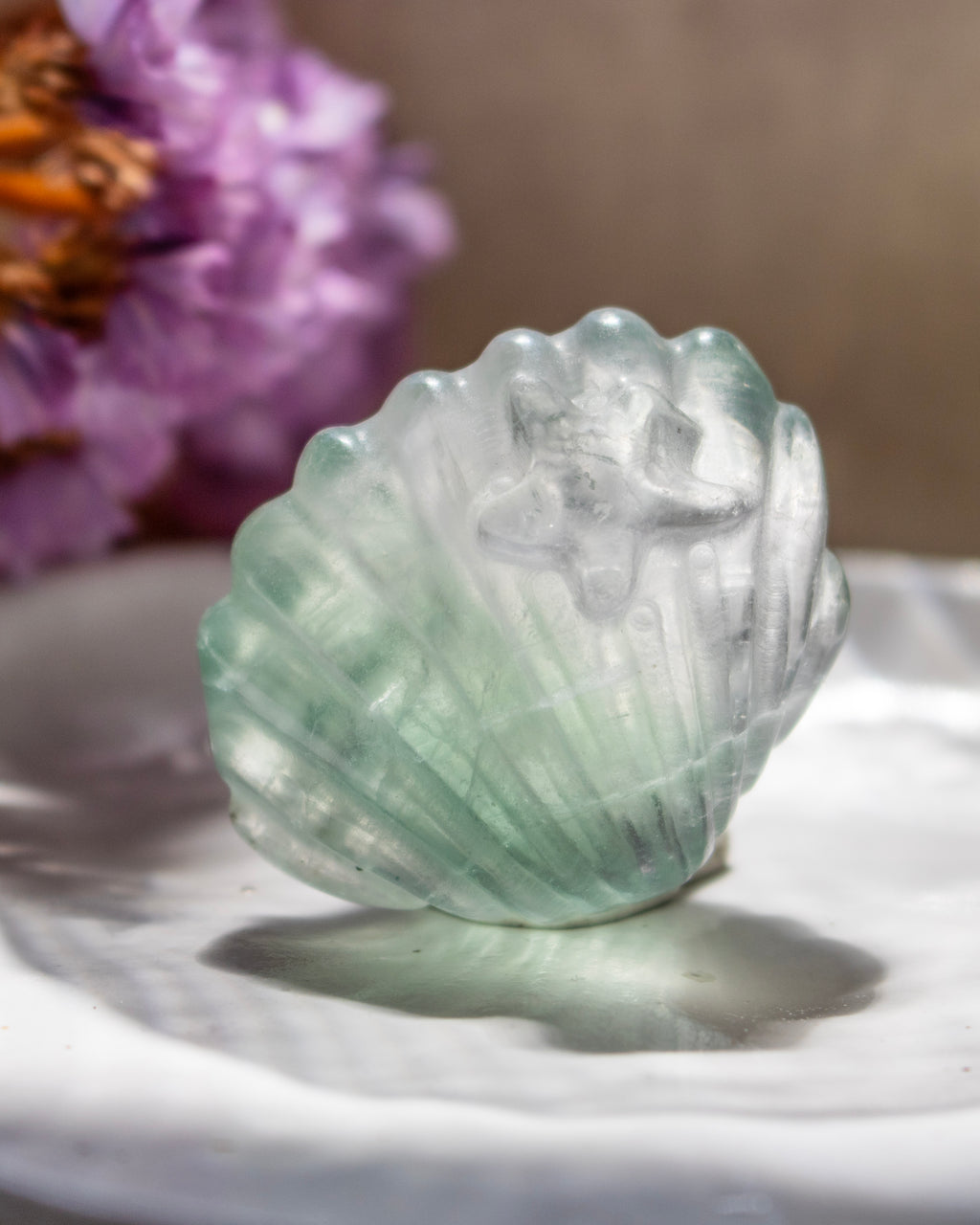 Greenish crystal-like object on a white surface with a blurred purple flower in the background