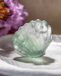 Greenish crystal-like object on a white surface with a blurred purple flower in the background