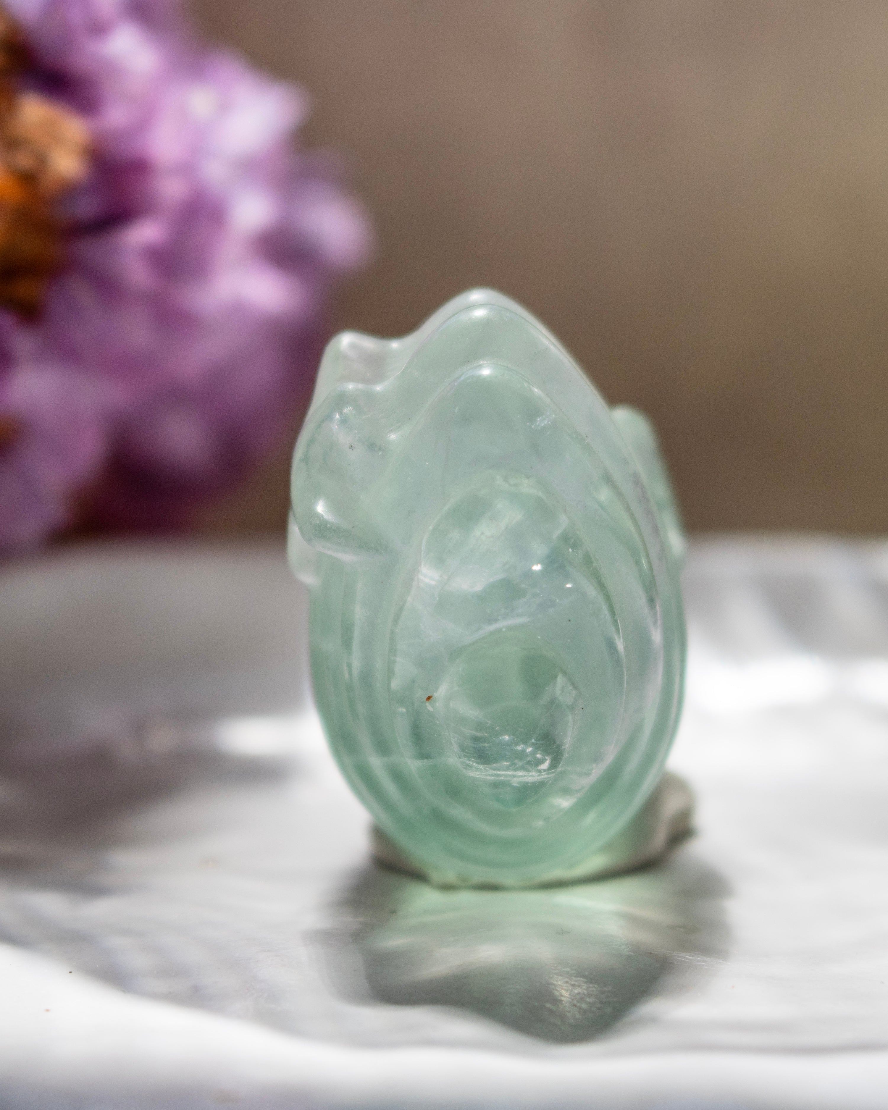 Green crystal carving on a marble surface with blurred purple flowers in the background