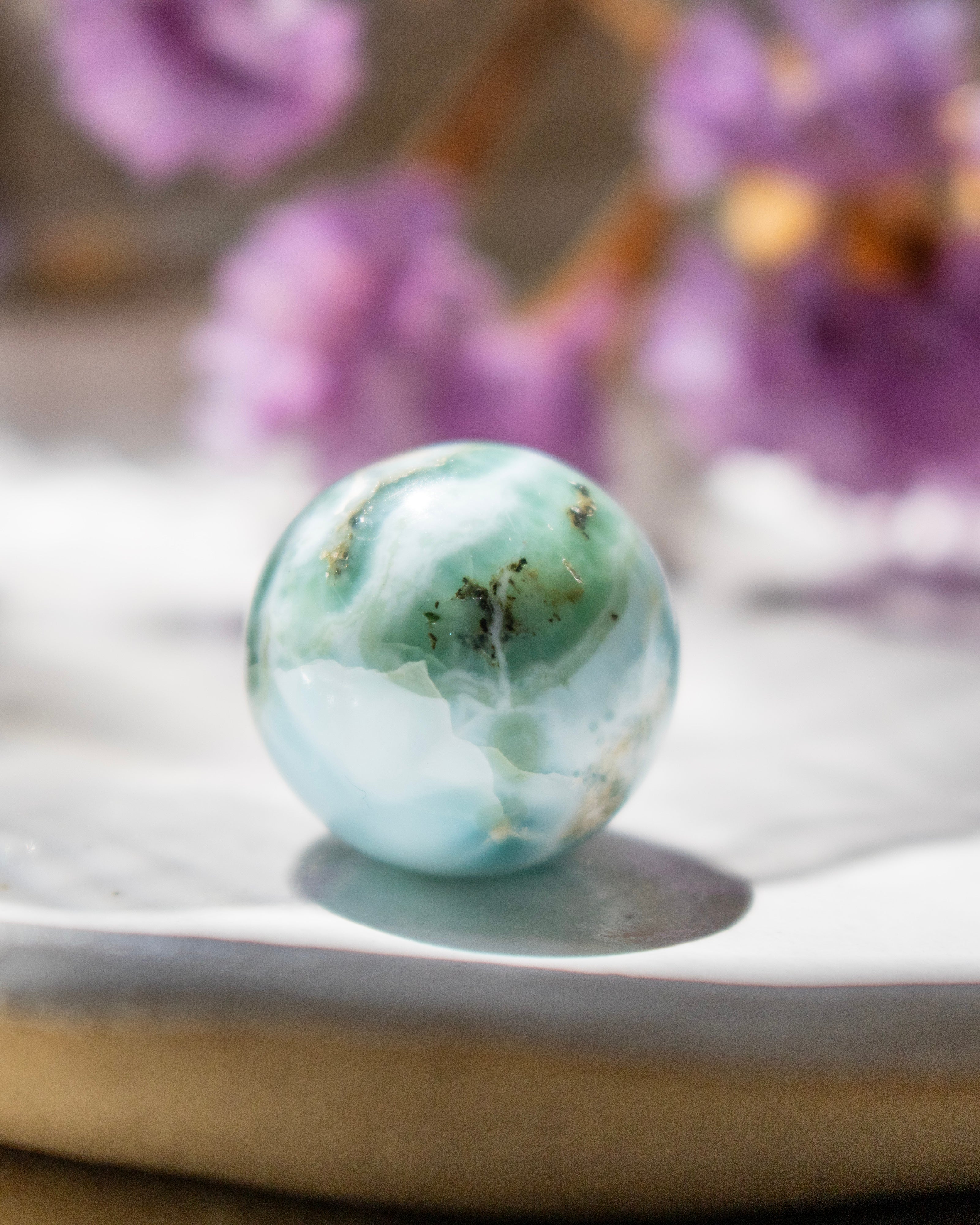 Turquoise marble-like sphere on a white surface with blurred purple flowers in the background