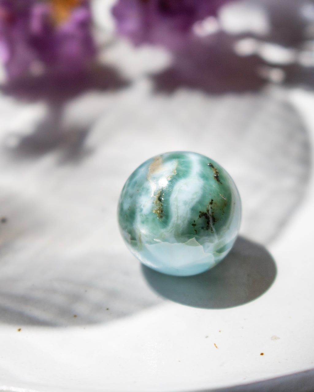 Marble-like sphere with green and white patterns on a white surface with blurred purple flowers in the background