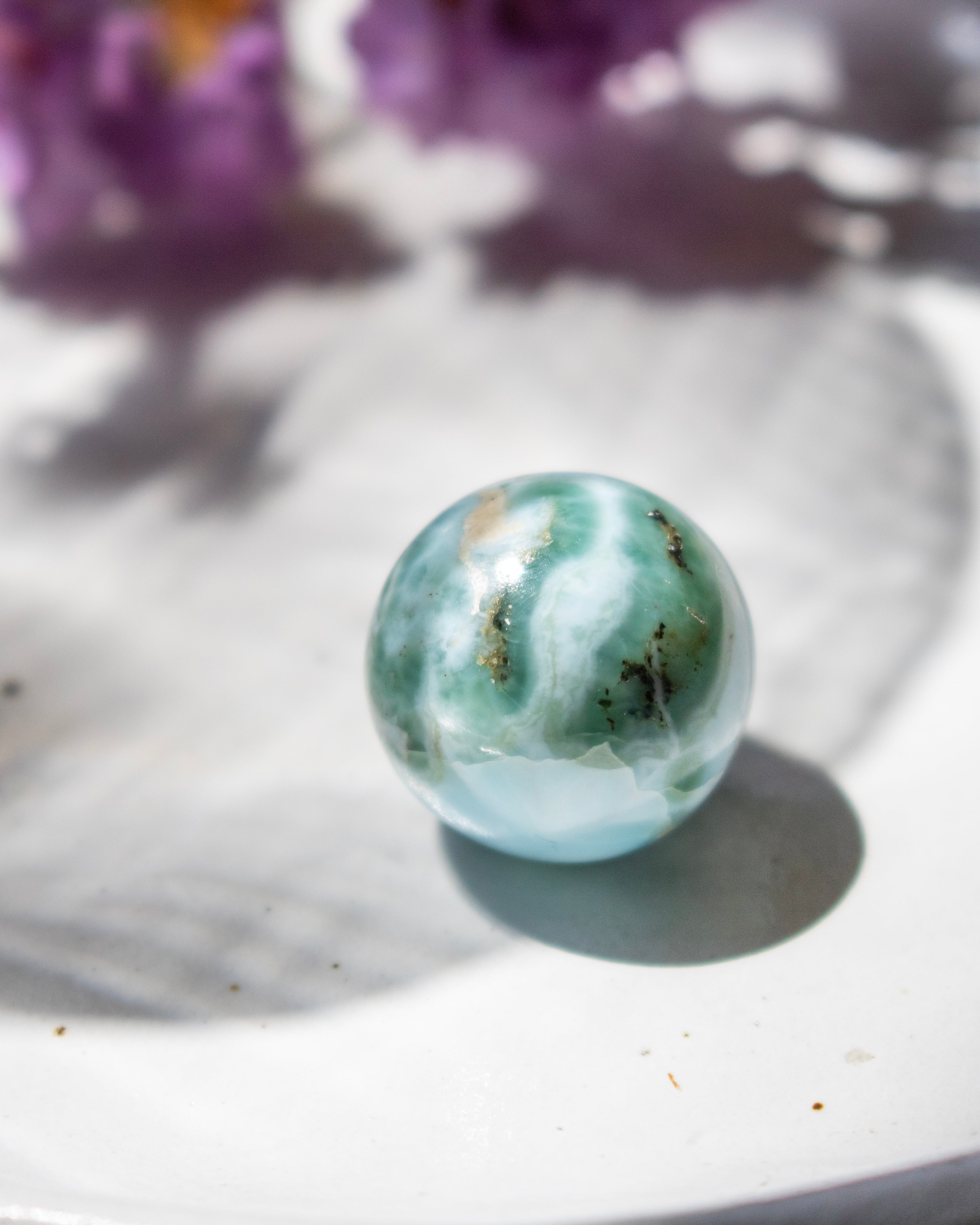Marble-like sphere with green and white patterns on a white surface with blurred purple flowers in the background