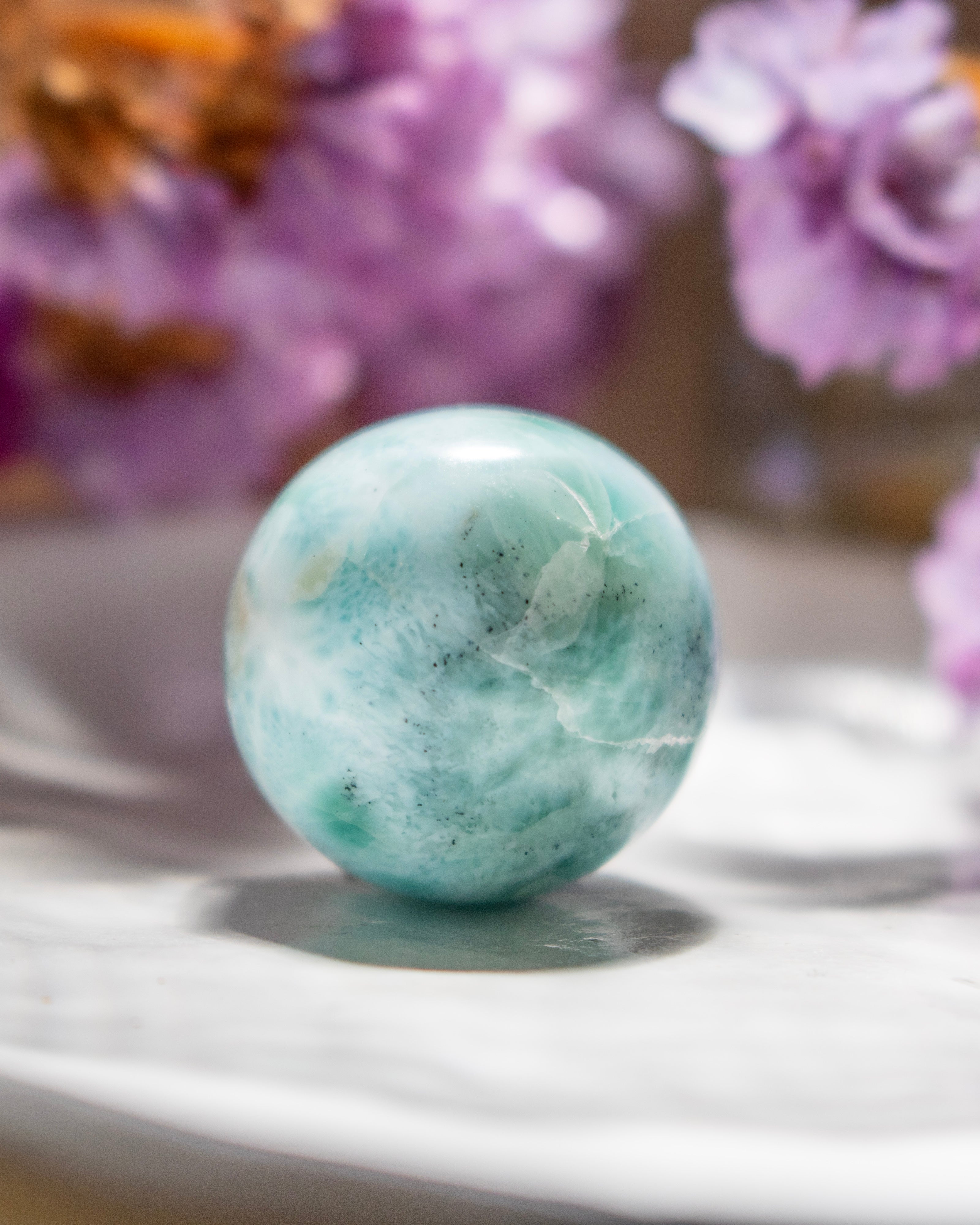 Turquoise marble sphere on a white surface with blurred purple flowers in the background