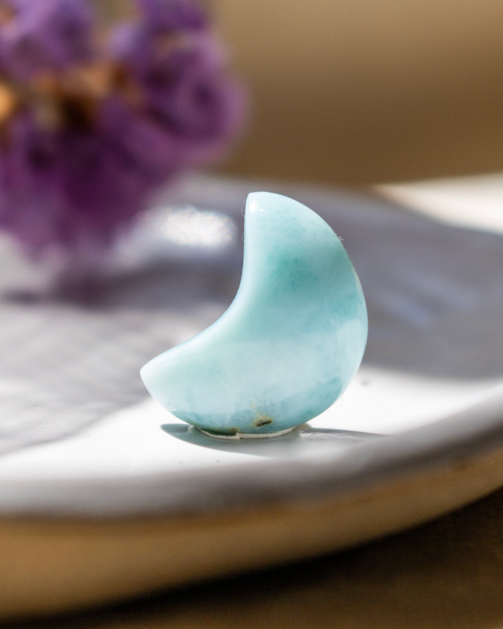 Teal crescent moon-shaped object on a white surface with blurred purple flowers in the background