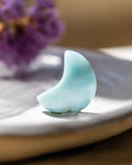 Teal crescent moon-shaped object on a white surface with blurred purple flowers in the background