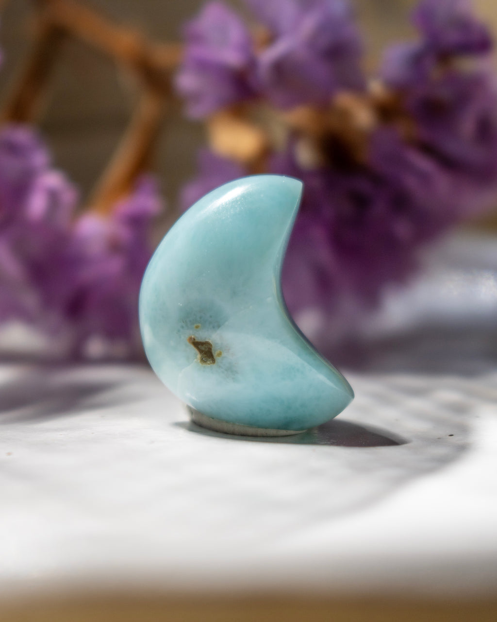 Teal stone shaped like a crescent moon on a white surface with blurred purple flowers in the background
