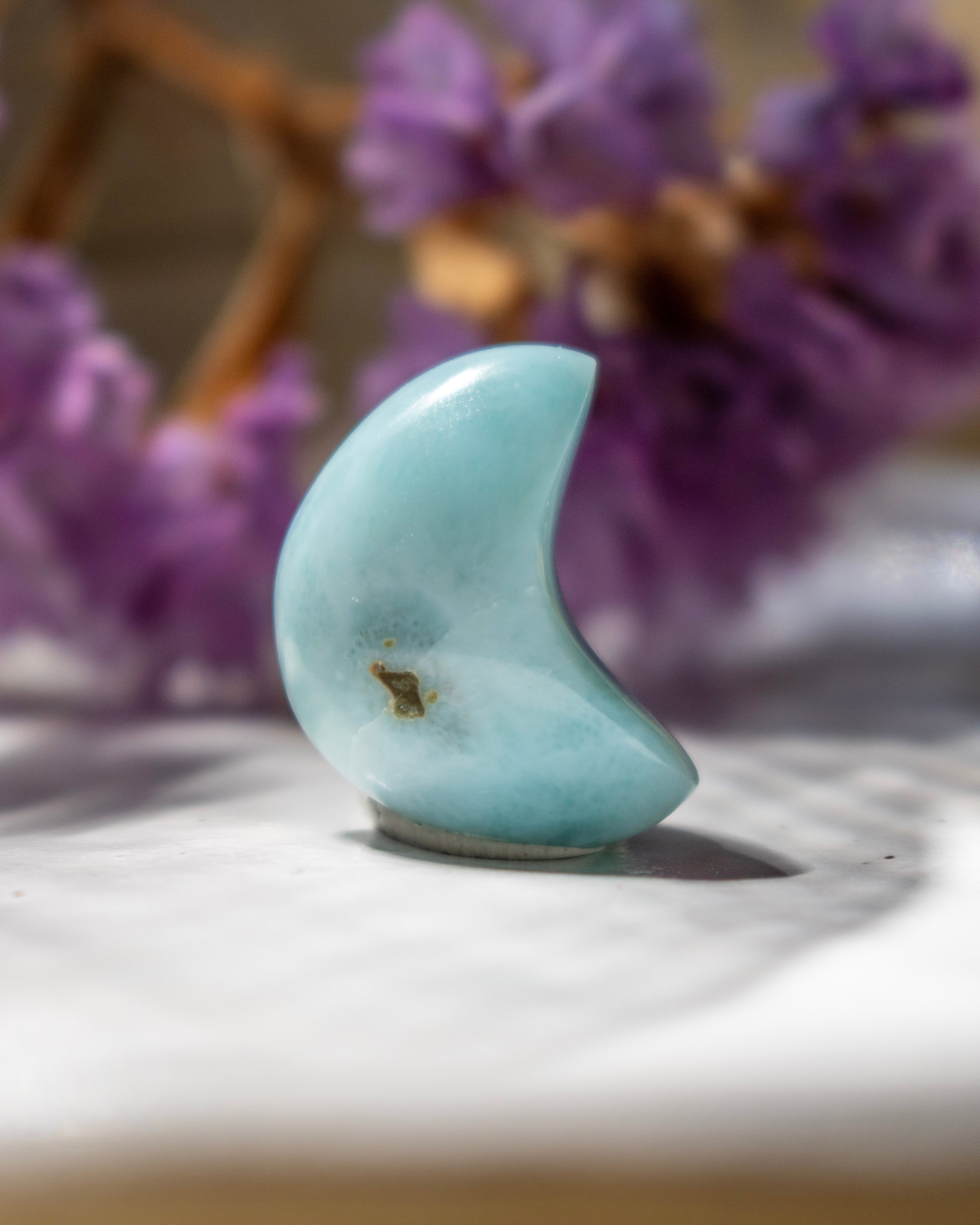 Teal stone shaped like a crescent moon on a white surface with blurred purple flowers in the background