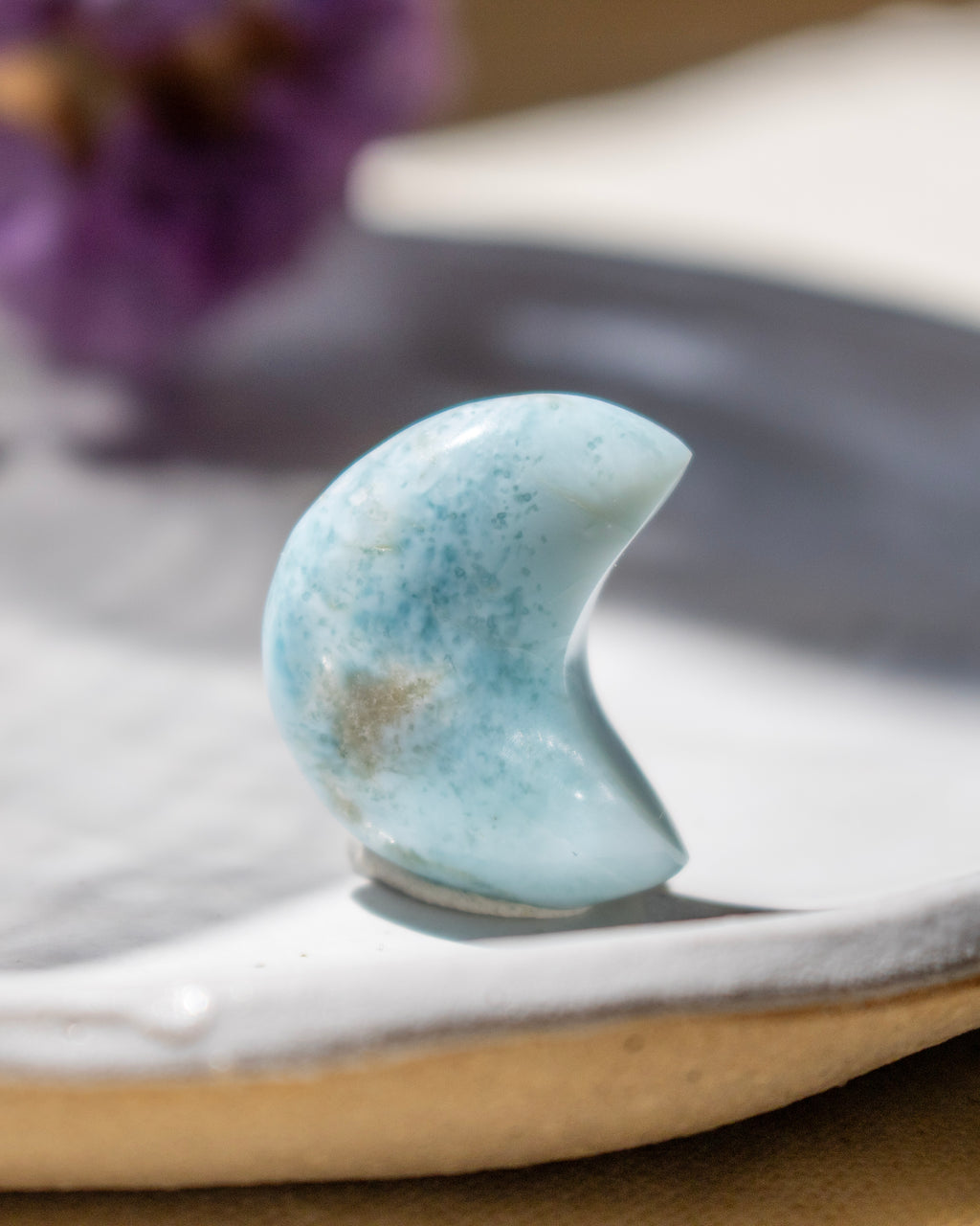 Blue stone in crescent shape on a white surface with blurred purple flowers in the background