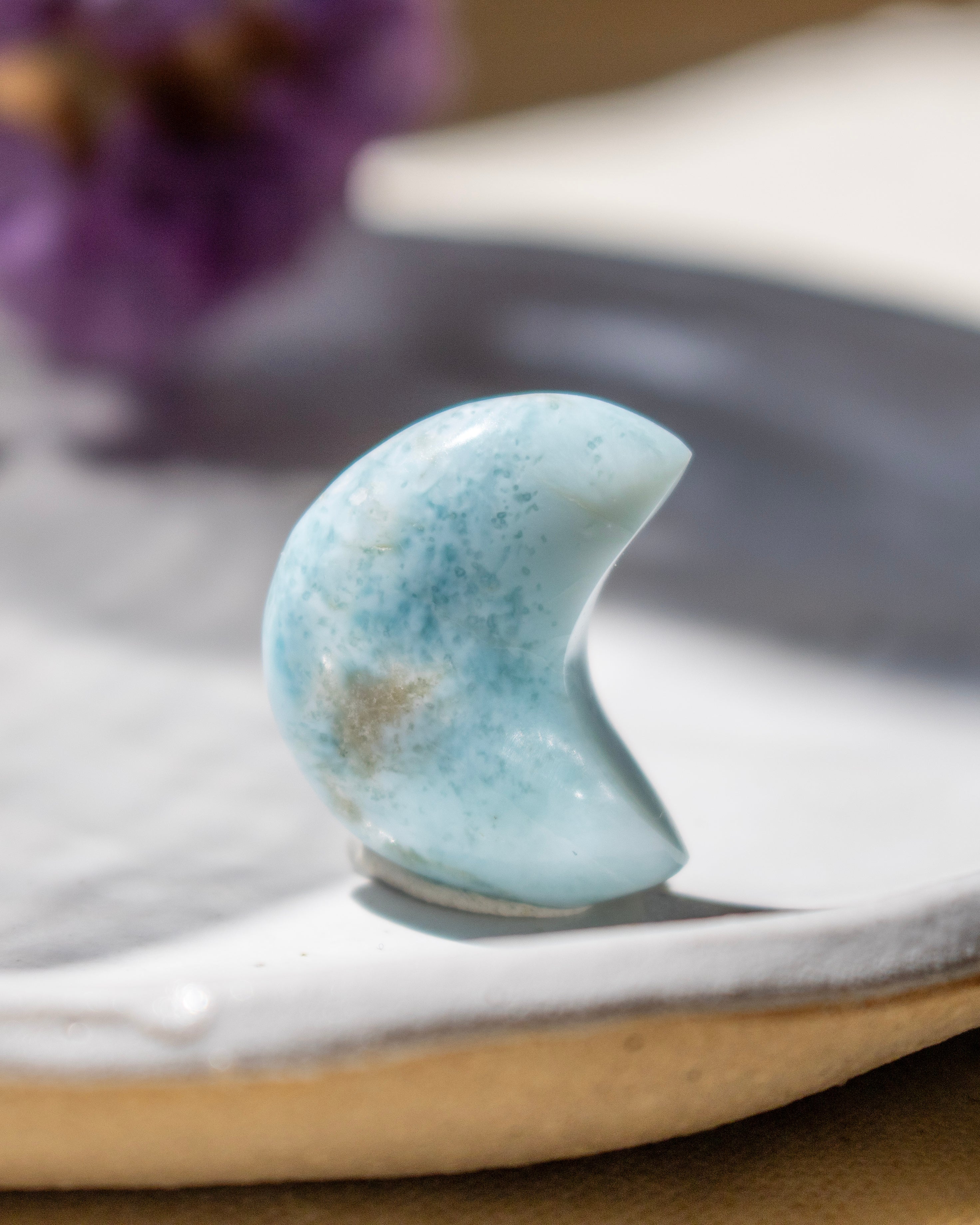 Blue stone in crescent shape on a white surface with blurred purple flowers in the background