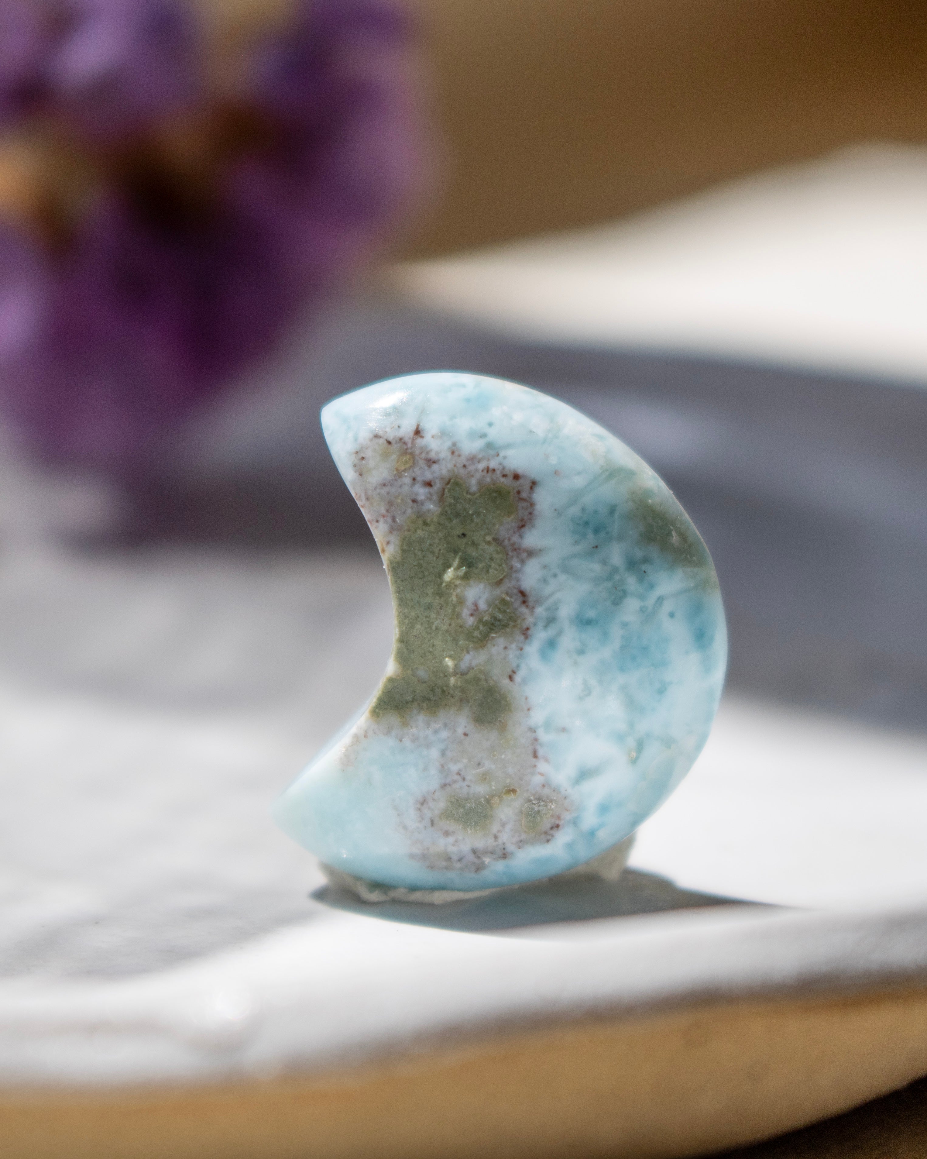 Larimar stone shaped like a crescent moon on a marble surface with a blurred purple flower in the background.