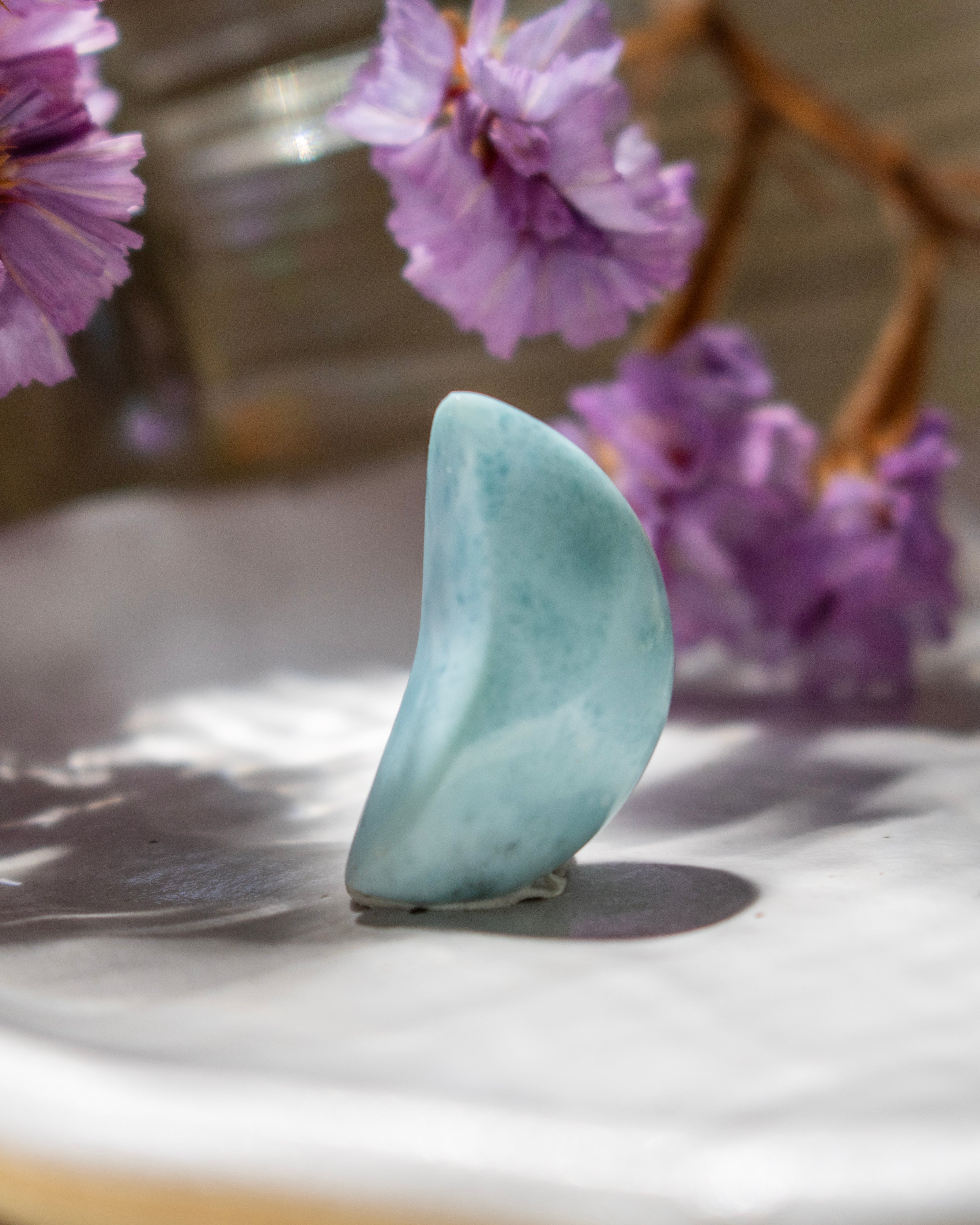 Teardrop-shaped light blue stone on a marble surface with purple flowers in the background
