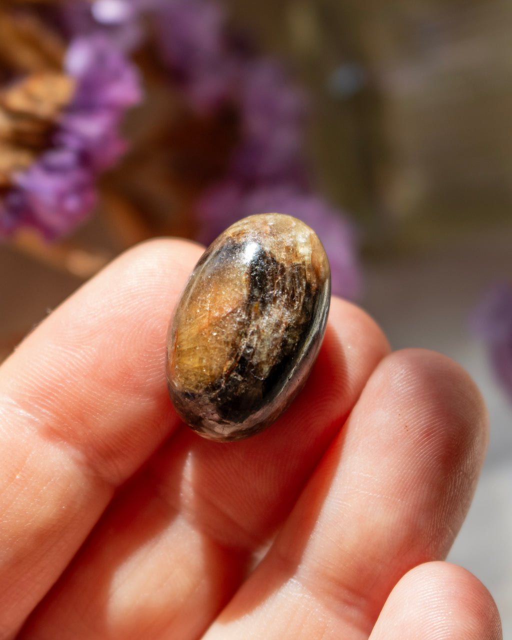 Hand holding a brown stone with blurred purple flowers in the background