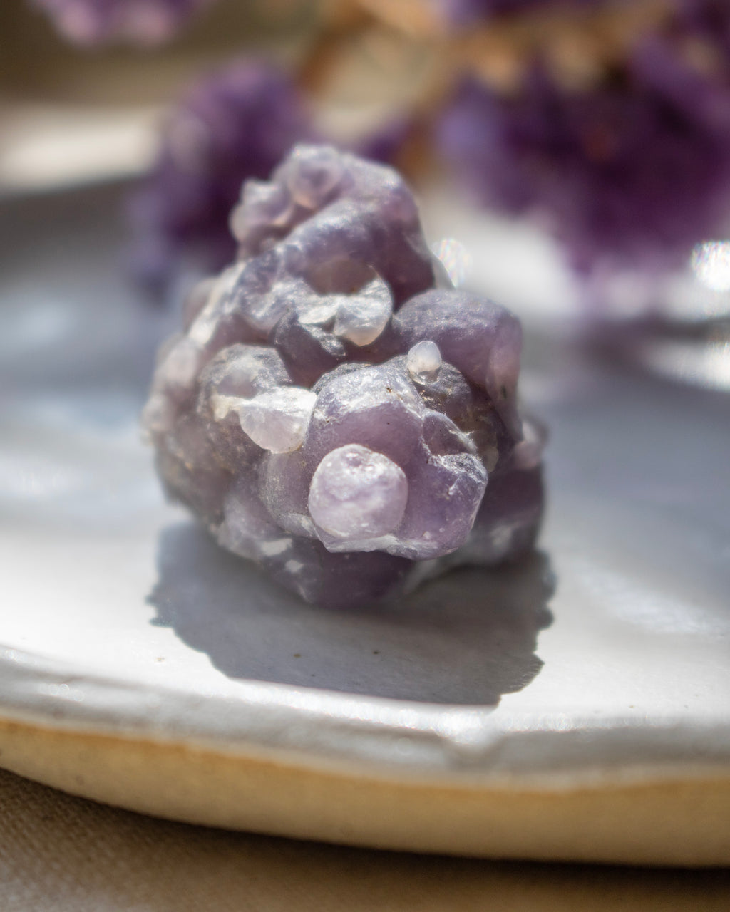 Close-up of a purple crystal on a reflective surface with blurred purple flowers in the background.