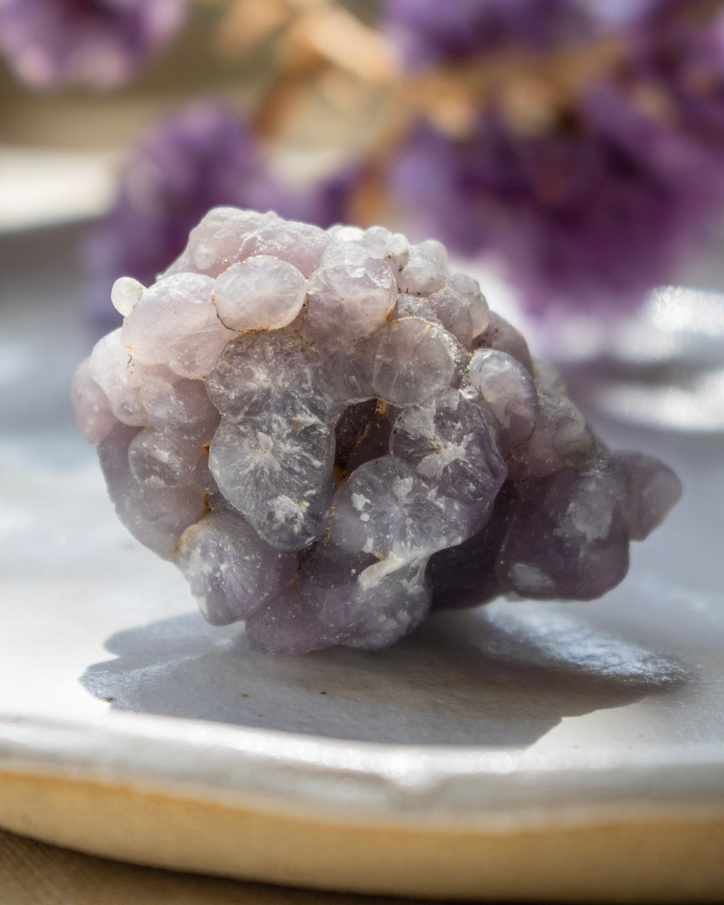 Close-up of a purple crystal on a reflective surface with blurred purple flowers in the background.