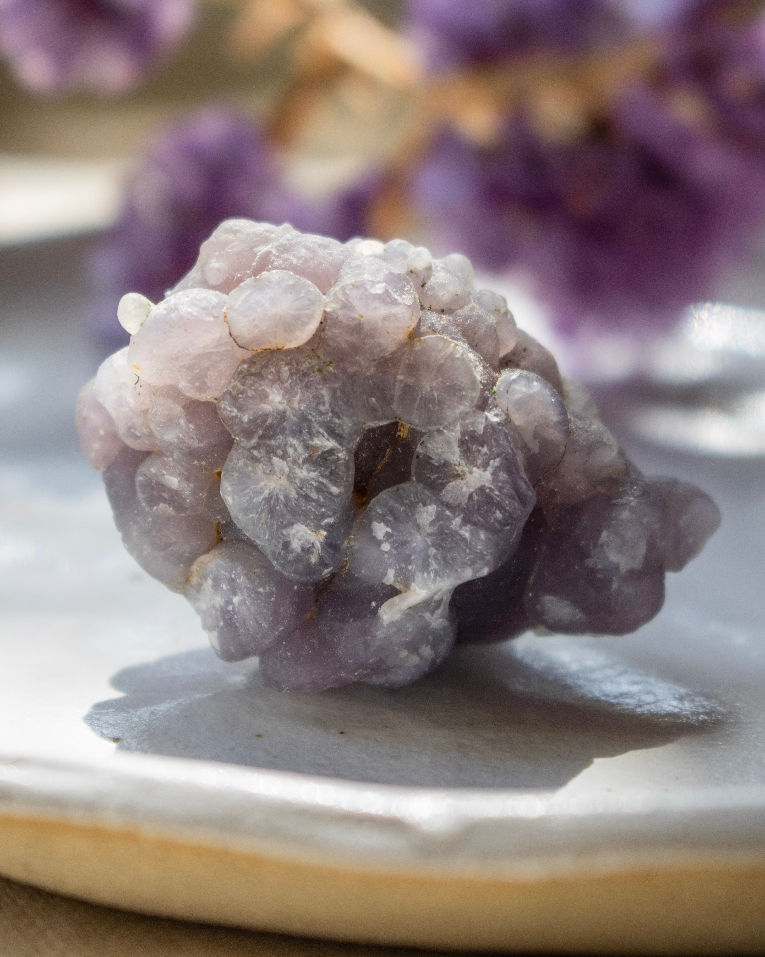 Close-up of a purple crystal on a reflective surface with blurred purple flowers in the background.