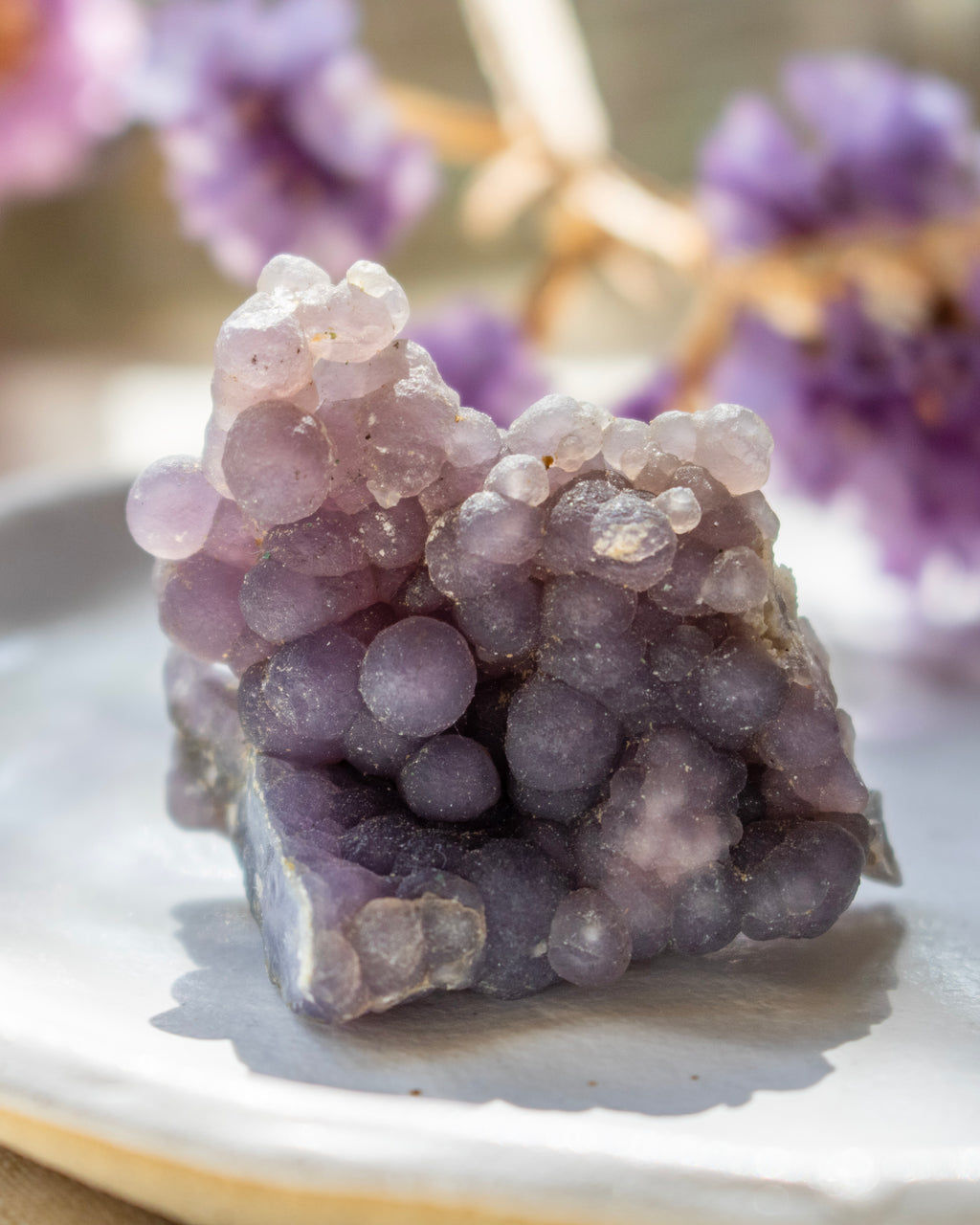 Close-up of a purple crystal on a reflective surface with blurred purple flowers in the background.