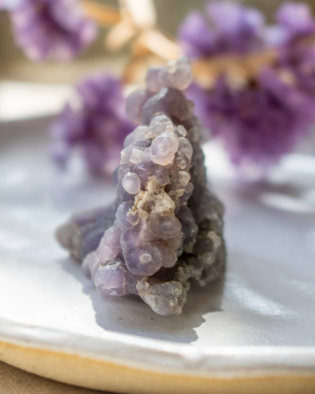 Close-up of a purple crystal on a reflective surface with blurred purple flowers in the background.