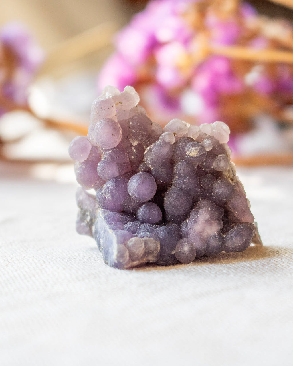 Purple crystal cluster on a white surface with blurred pink flowers in the background