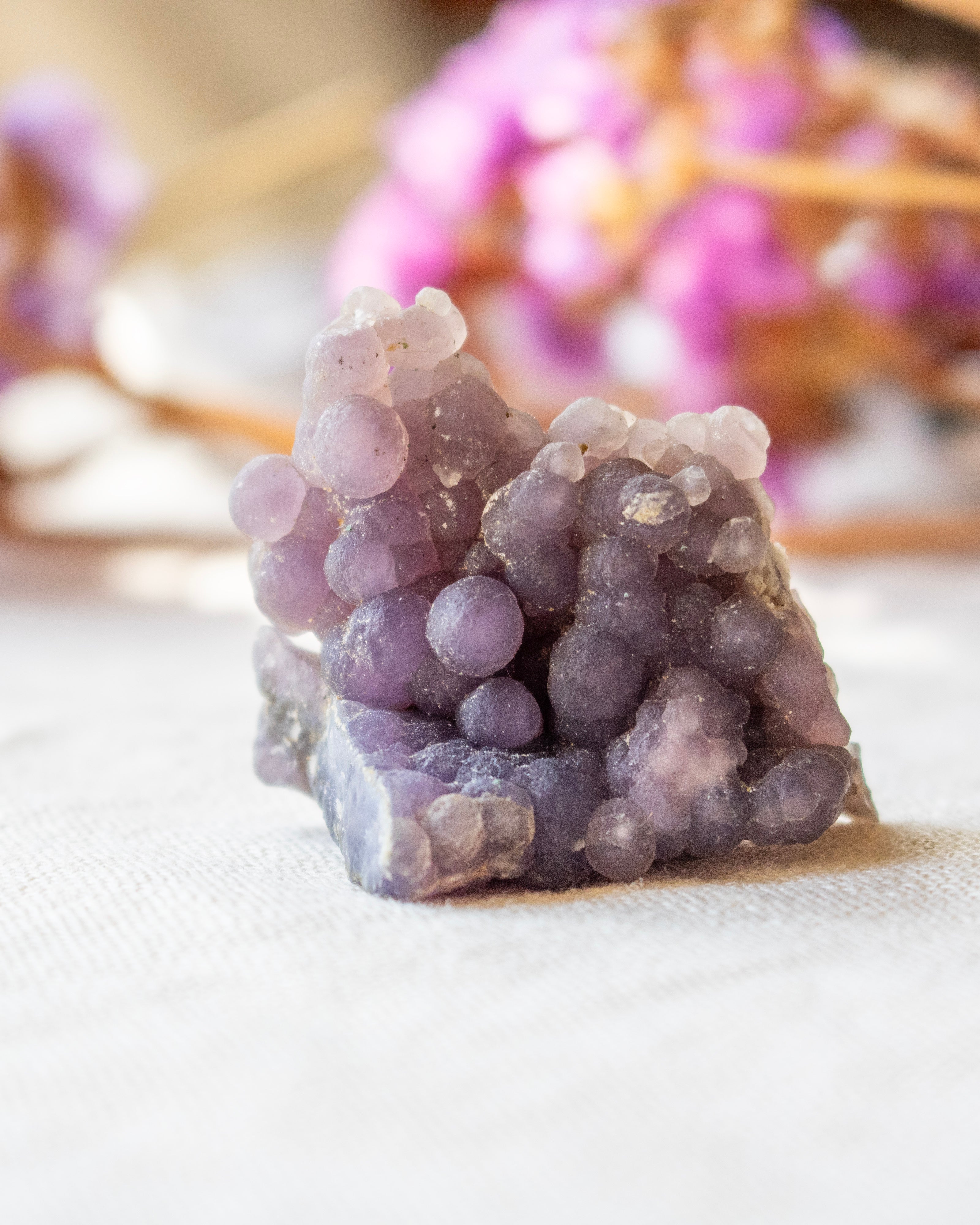 Purple crystal cluster on a white surface with blurred pink flowers in the background