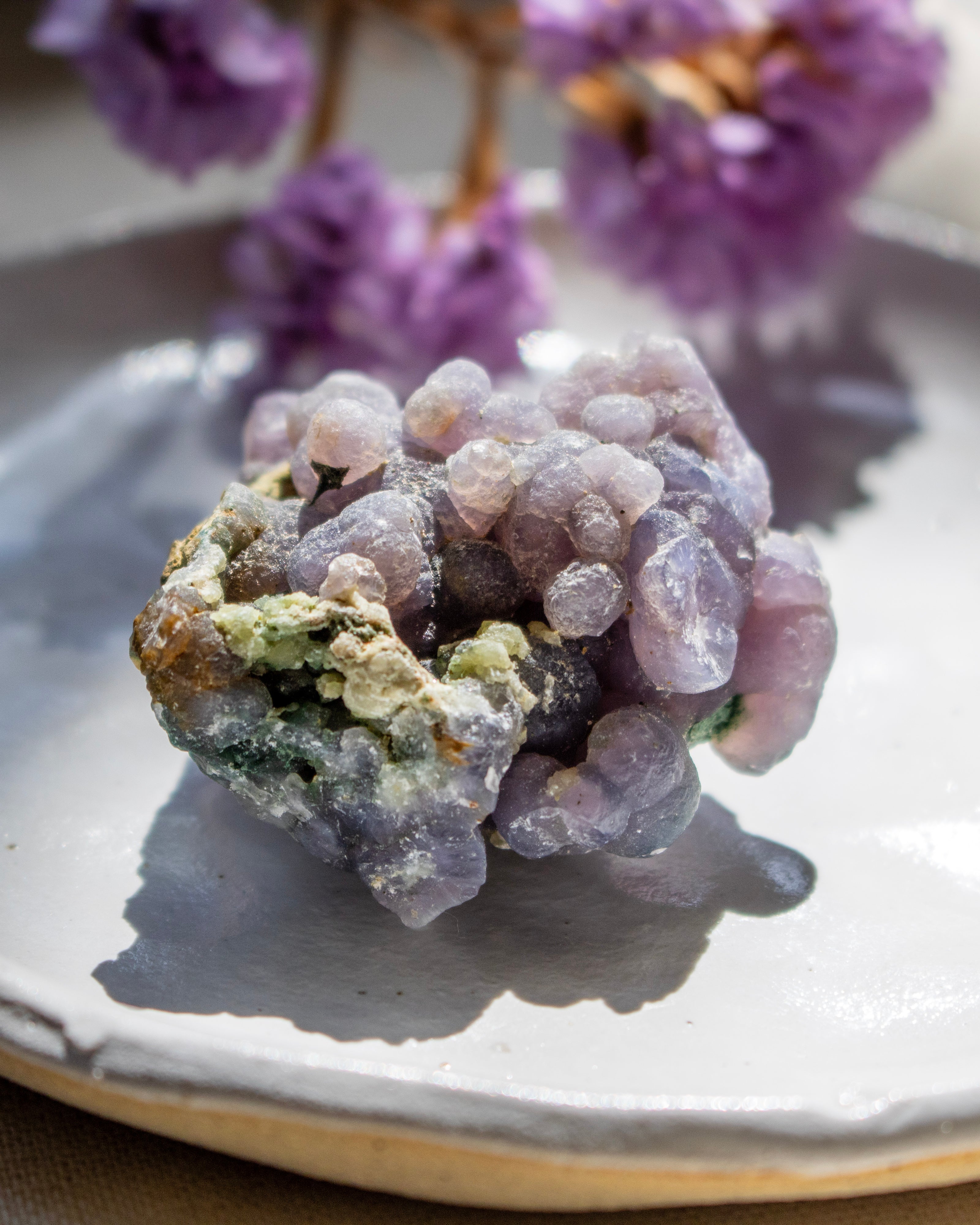 Close-up of a purple crystal on a reflective surface with blurred purple flowers in the background.