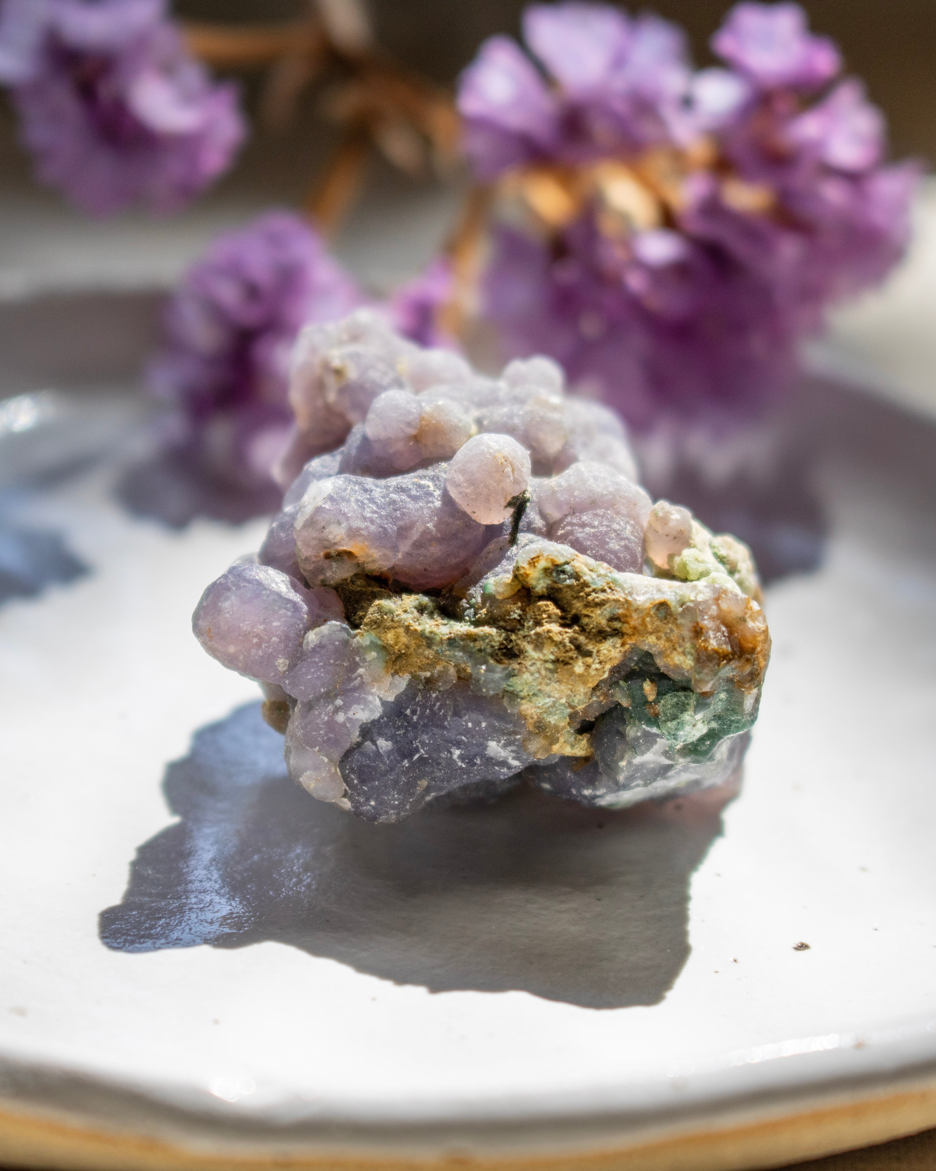 Close-up of a purple crystal on a reflective surface with blurred purple flowers in the background.