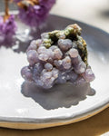 Close-up of a purple crystal on a reflective surface with blurred purple flowers in the background.
