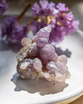 Purple crystal cluster on a white plate with purple flowers in the background