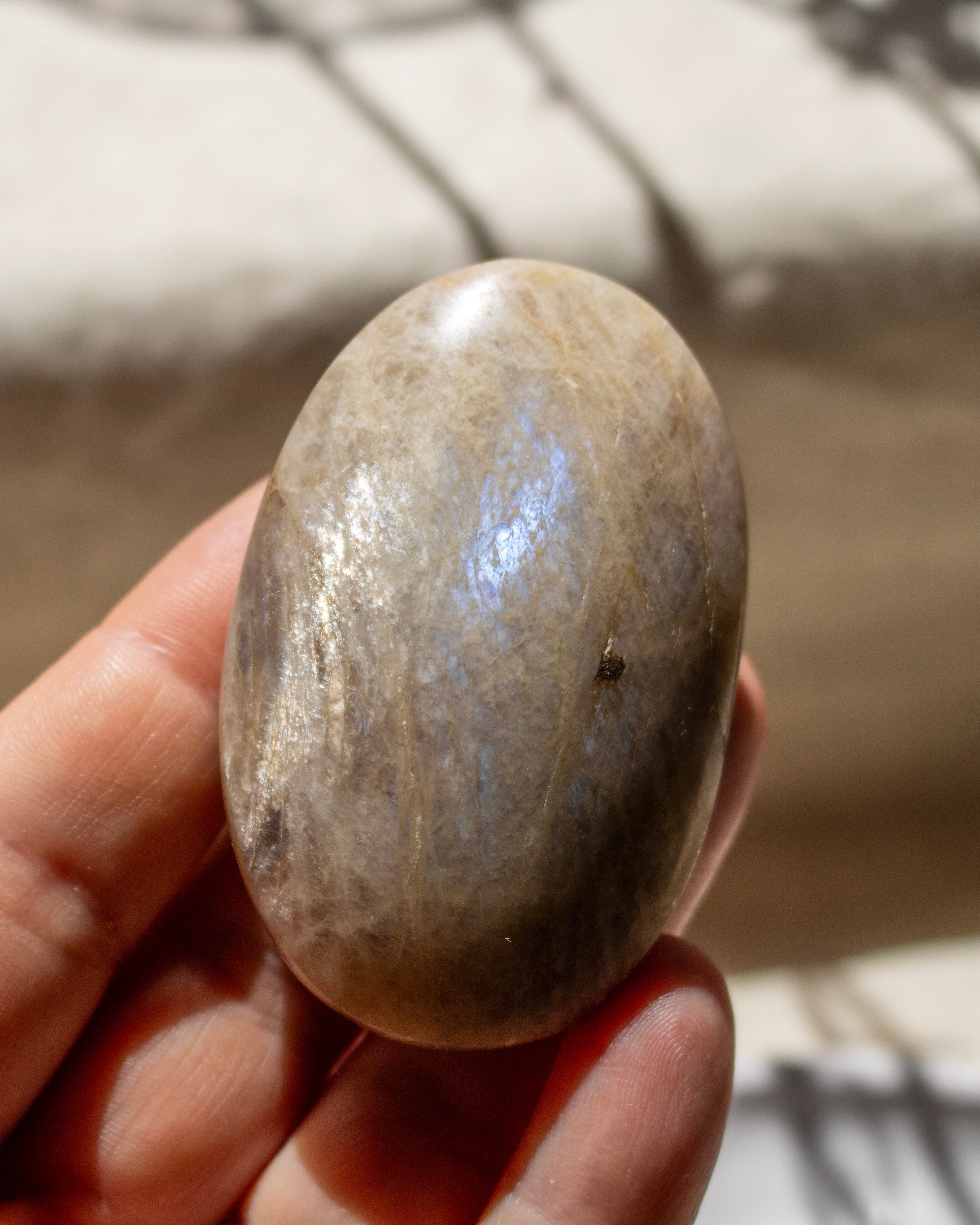A hand holding a polished oval stone with a smooth, matte finish and subtle gray-brown veining, set against a softly blurred background.