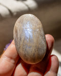 A polished oval stone with a smooth, slightly iridescent surface, held in a person's hand against a softly blurred neutral background.