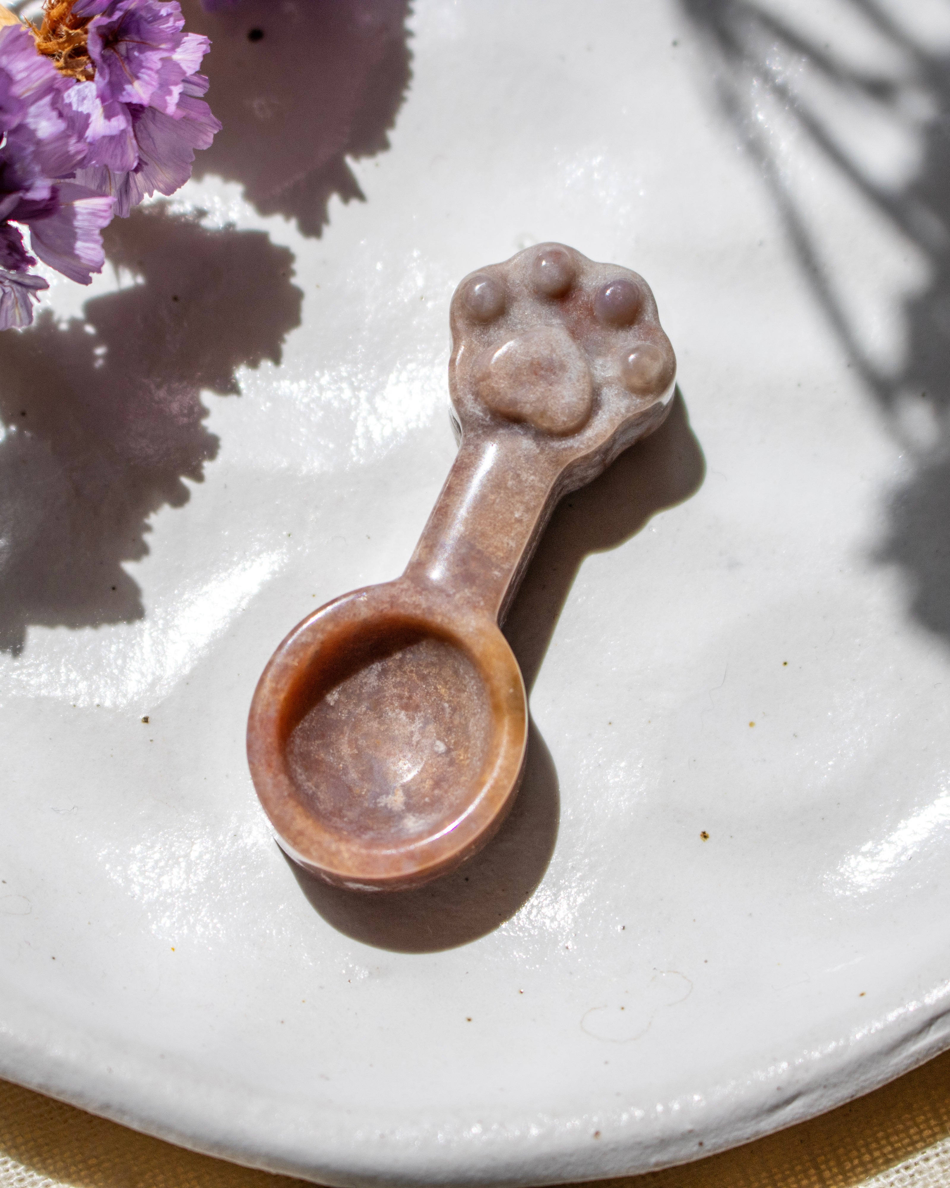 A small carved stone spoon with a paw-print handle, resting on a textured white ceramic plate with purple flowers in the corner.