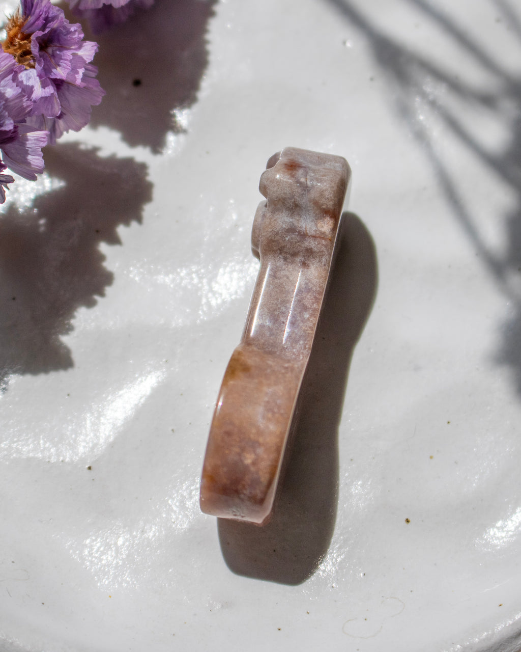 A polished carved stone figurine with a curved, handle-like shape, featuring natural brown and beige marbling, resting on a white textured surface with purple flowers in the corner.
