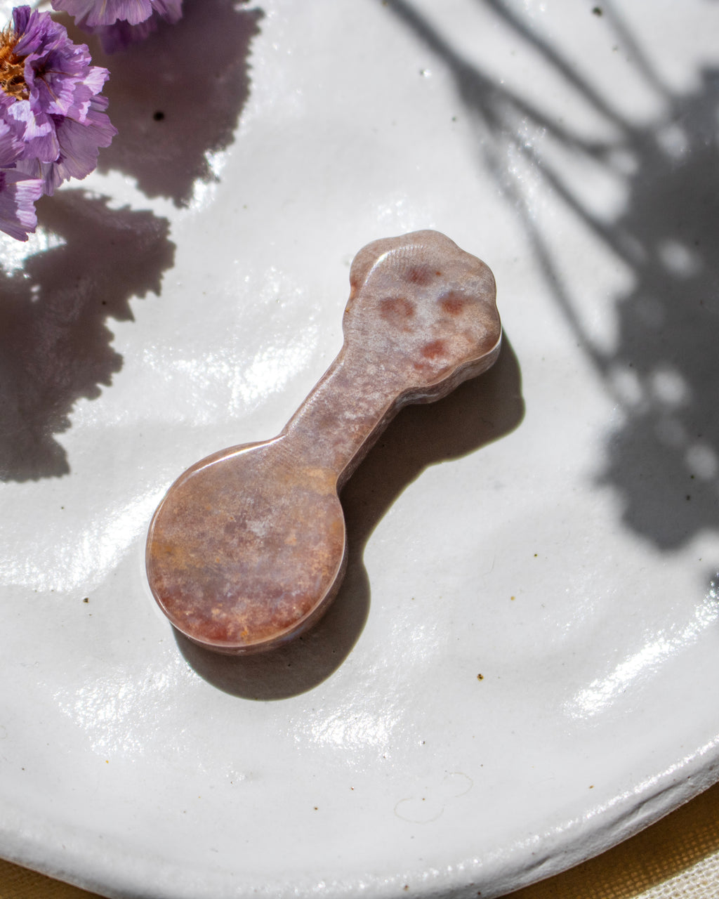 A small spoon-shaped decorative object with a rounded handle, made of marbled stone in pink, peach, and brown tones, resting on a white ceramic plate with purple flowers in the corner.