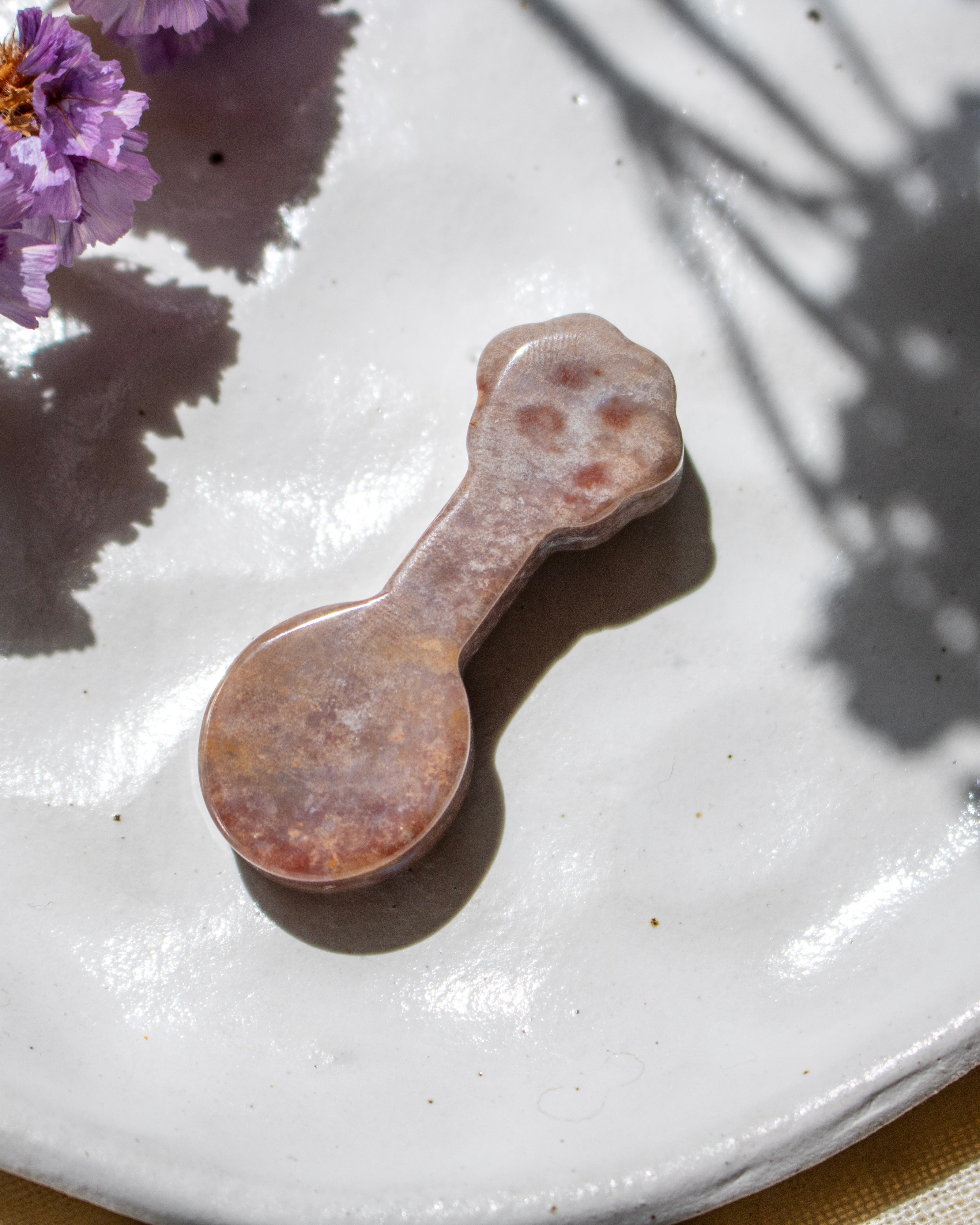 A small spoon-shaped decorative object with a rounded handle, made of marbled stone in pink, peach, and brown tones, resting on a white ceramic plate with purple flowers in the corner.