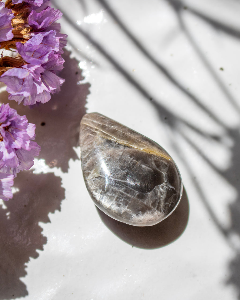A polished teardrop-shaped clear quartz crystal with natural inclusions, resting on a white textured surface with purple flowers and palm leaf shadows.