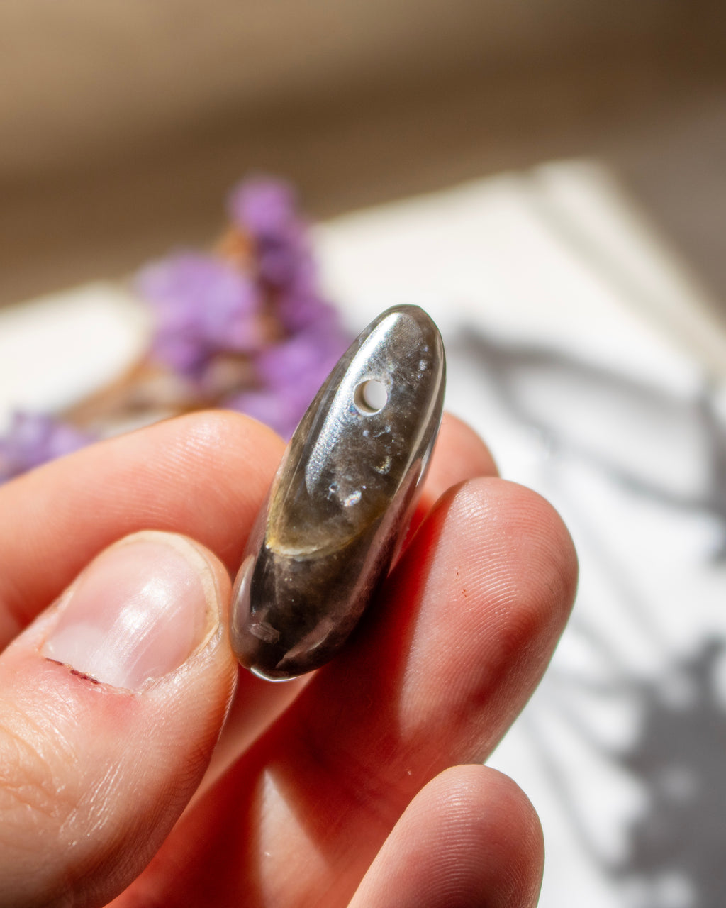 A polished dark gray stone bead with a small hole, held between fingers against a soft-focus background with purple flowers.