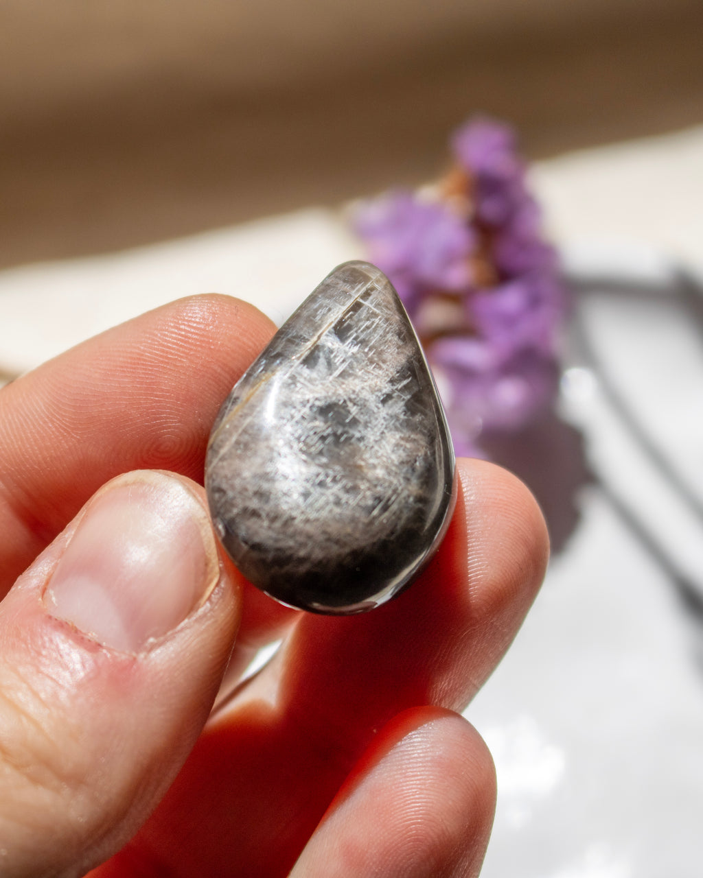 A hand holding a polished teardrop-shaped gray stone with visible crystalline internal structure, photographed close-up against a softly blurred background with purple flowers.