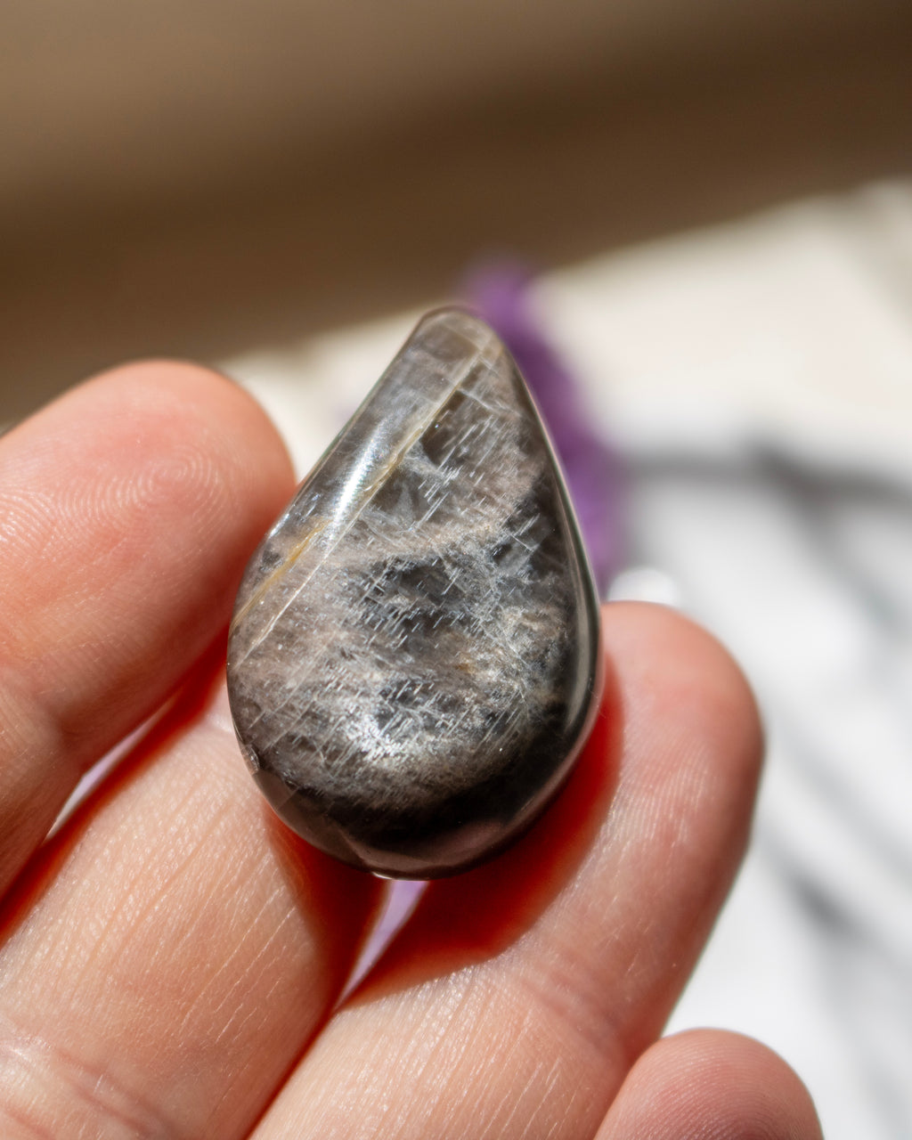 A polished teardrop-shaped tumbled stone with a metallic silver-gray sheen and fine linear texture, held between fingers to show its size.