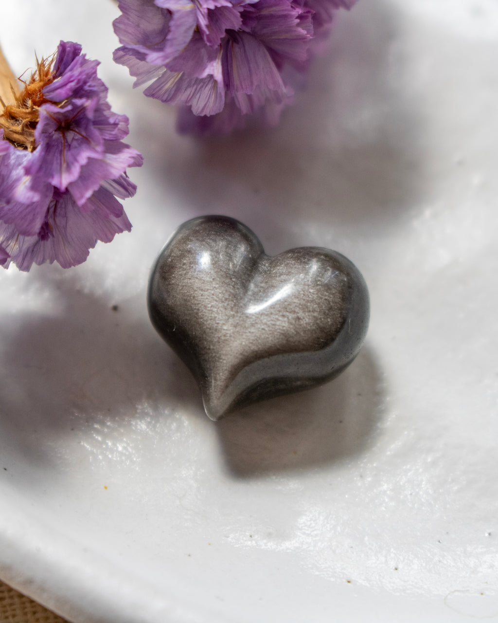 a black and silver heart shape stone on a white plate with soft shadows and purple flowers in the top left corner.