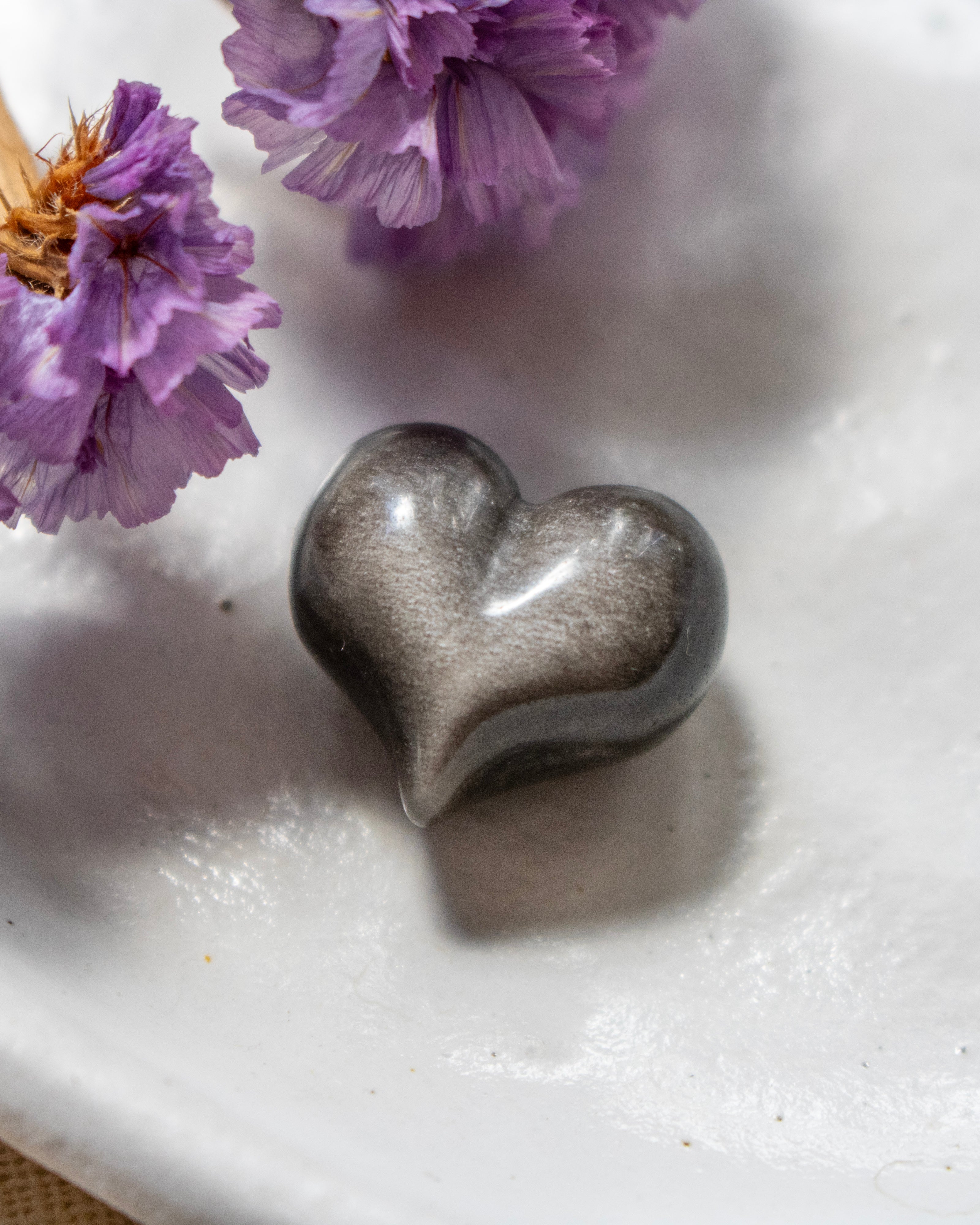 a black and silver heart shape stone on a white plate with soft shadows and purple flowers in the top left corner.