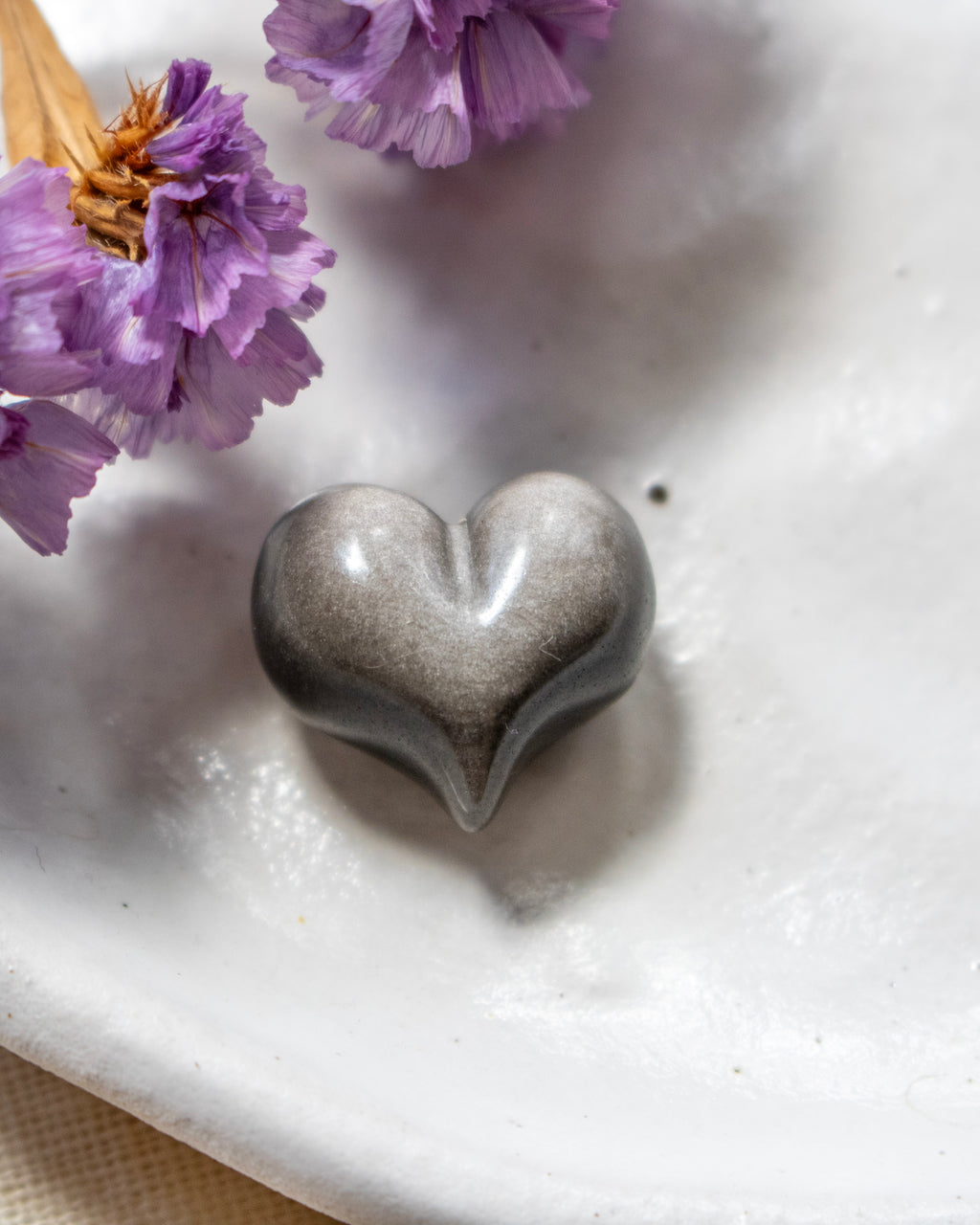 a black and silver heart shape stone on a white plate with soft shadows and purple flowers in the top left corner.