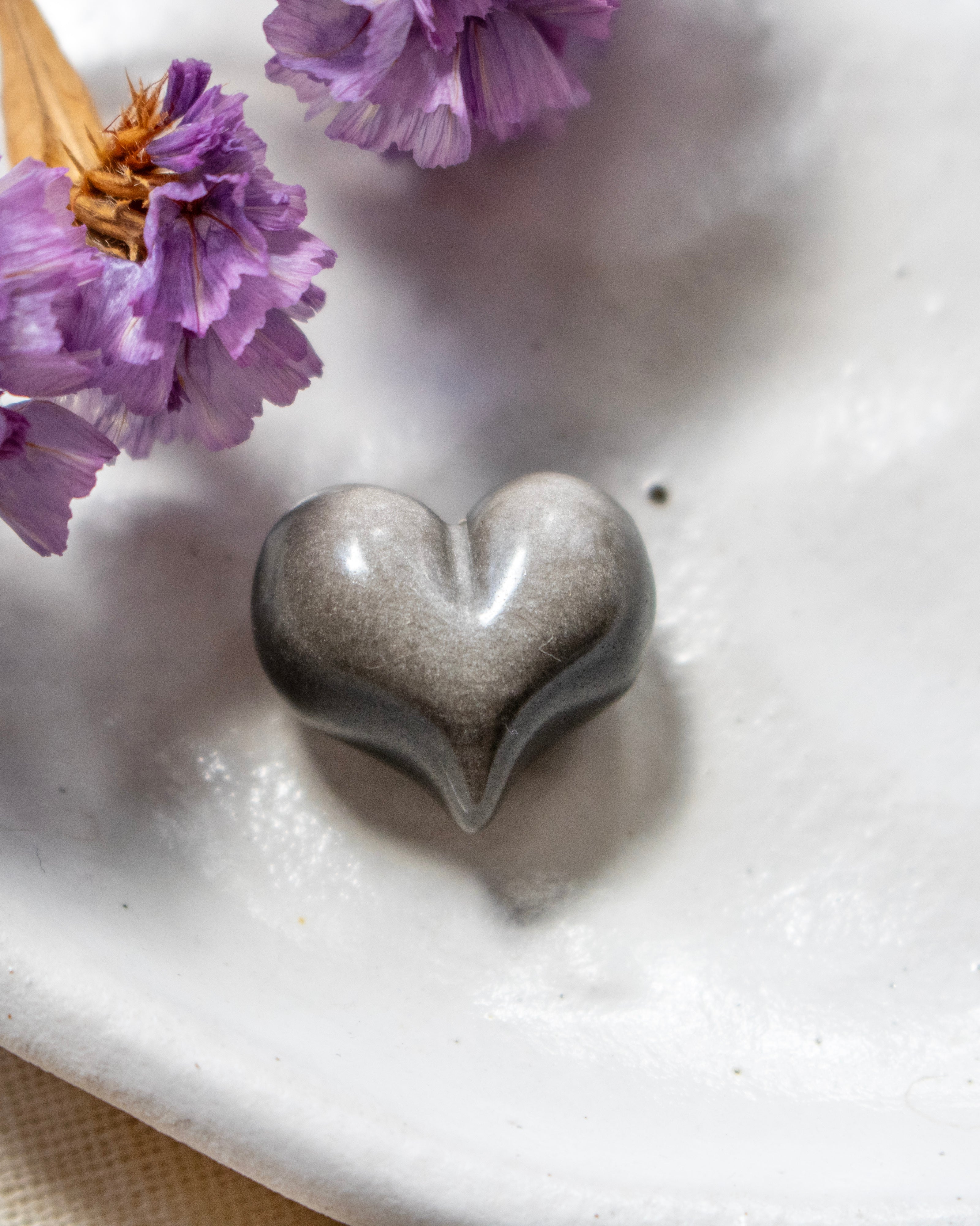 a black and silver heart shape stone on a white plate with soft shadows and purple flowers in the top left corner.