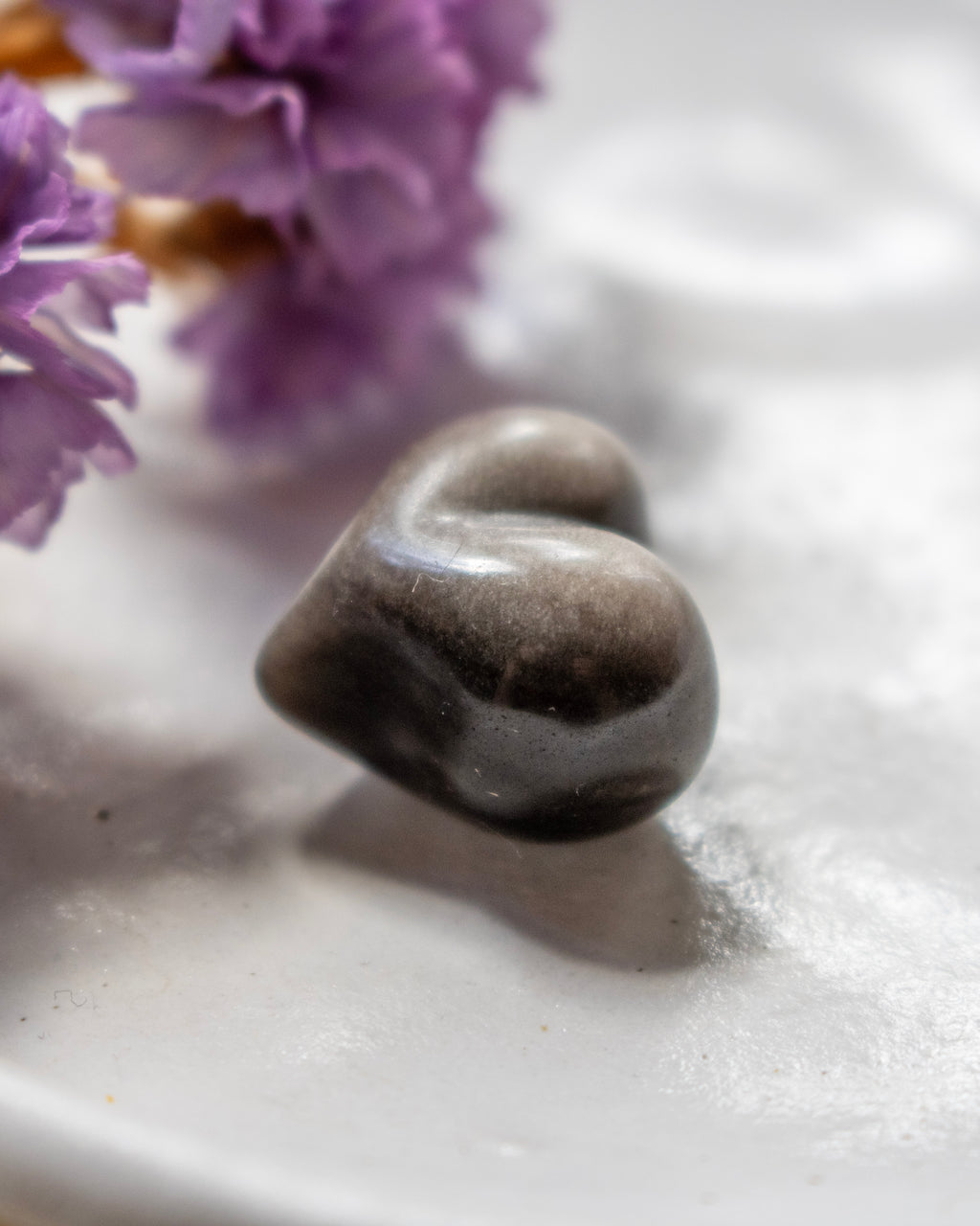 a black and silver heart shape stone on a white plate with soft shadows and purple flowers in the top left corner.